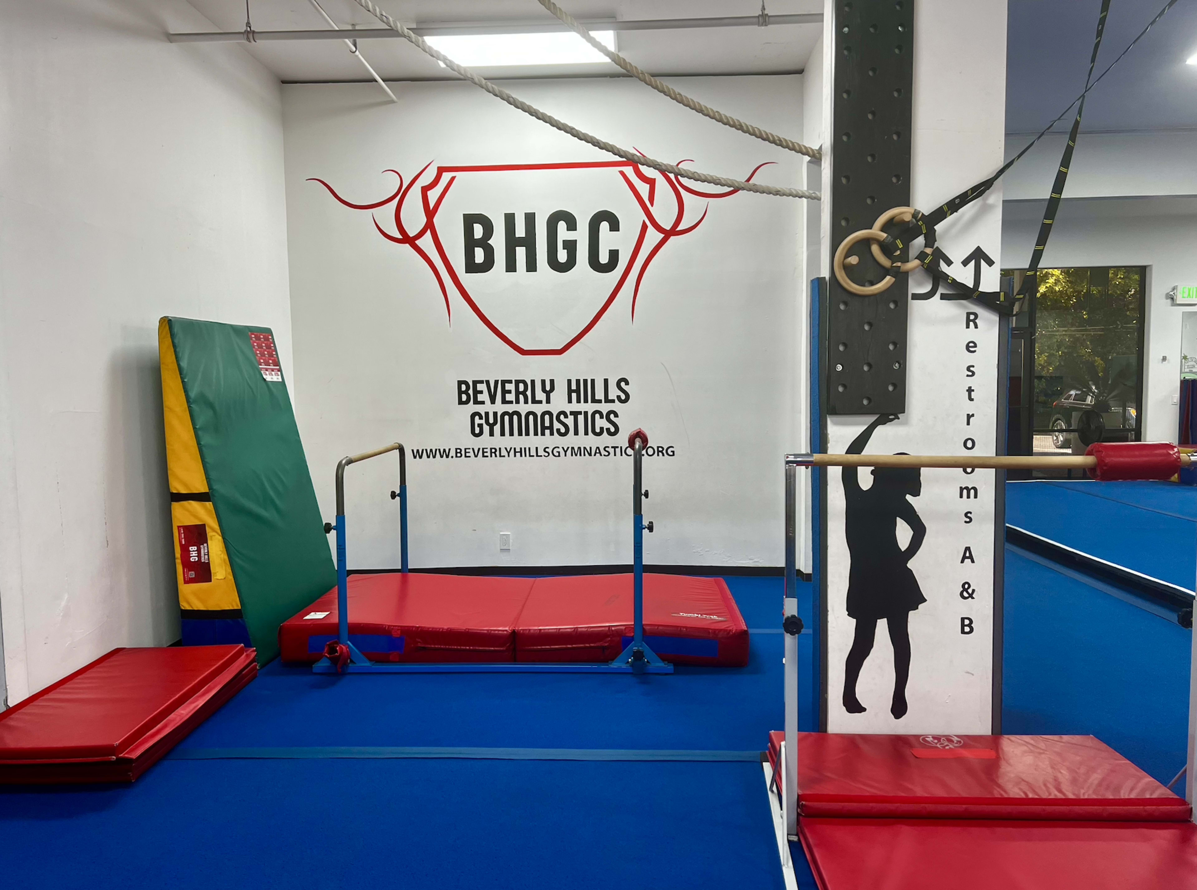 A gymnastics training area at Beverly Hills Gymnastics, featuring various gymnastic equipment, including a balance beam and rings, against a blue floor.