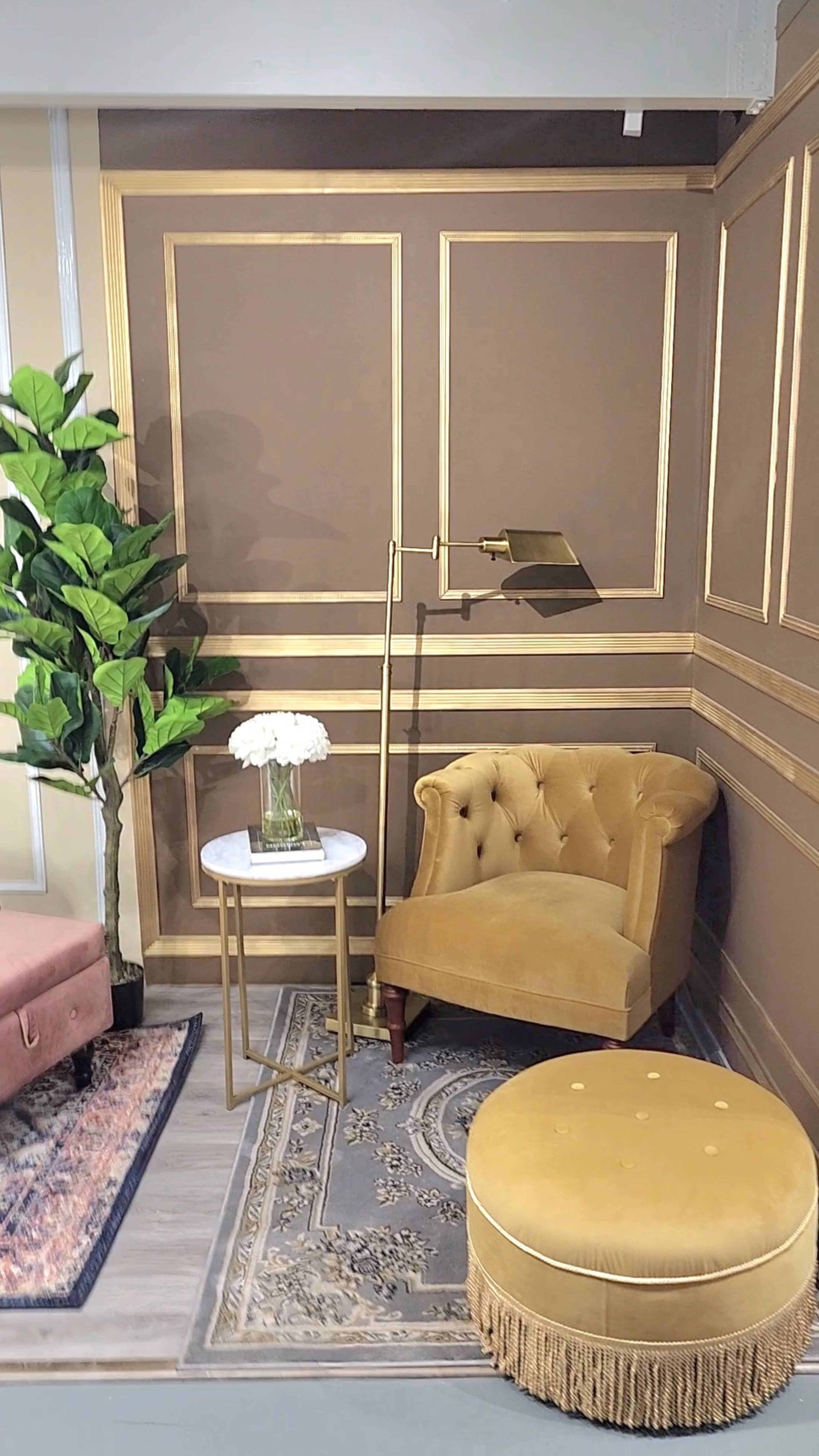 A corner of a room featuring a gold-accented wall, a tufted yellow chair, a small round table with flowers, a fringed ottoman, and a potted plant.