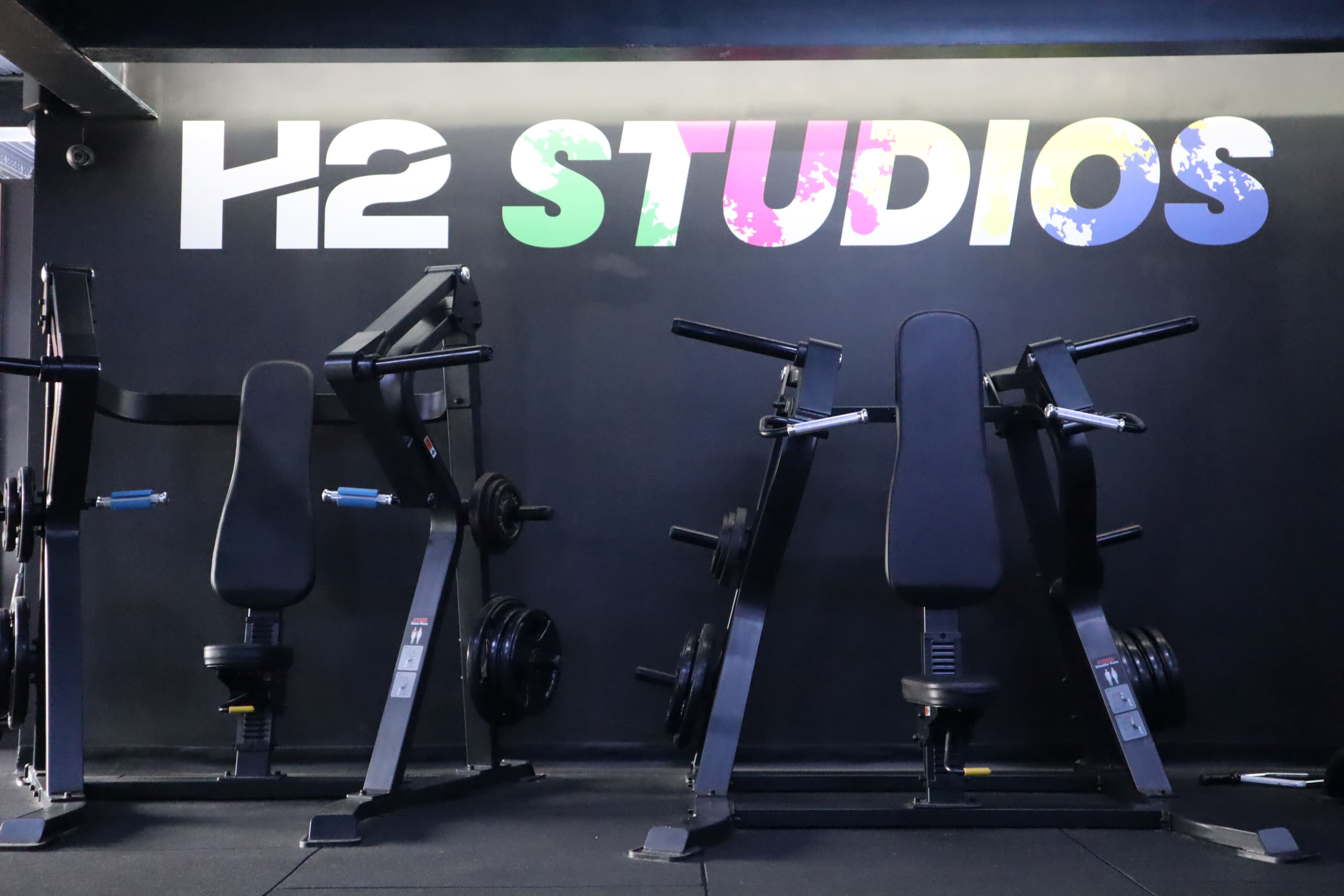 The image shows exercise equipment arranged in front of a black wall featuring the colorful text "H2 STUDIOS."