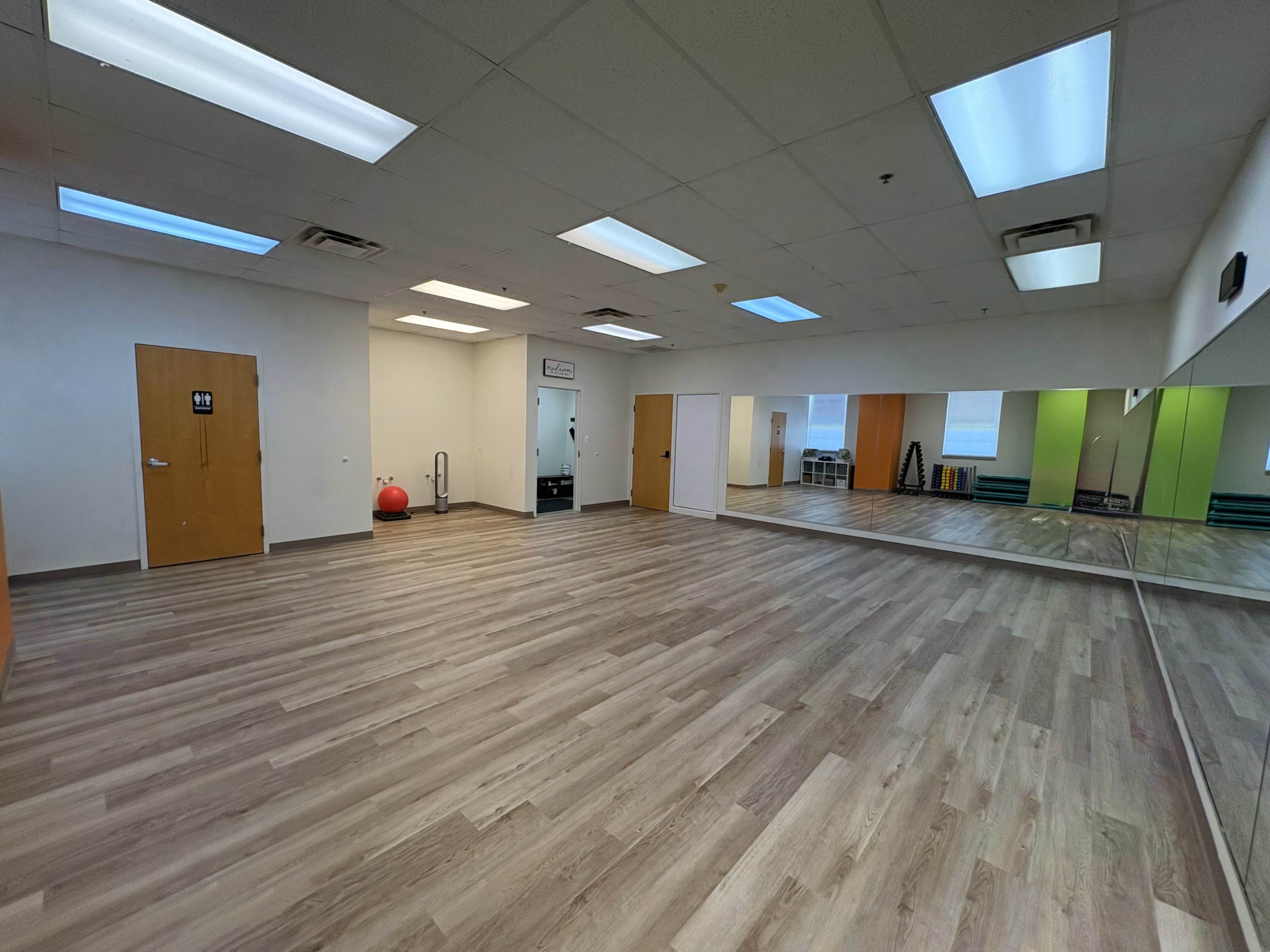 The image shows an empty gym studio with wooden flooring, mirrors on one wall, and a variety of exercise equipment in the background.