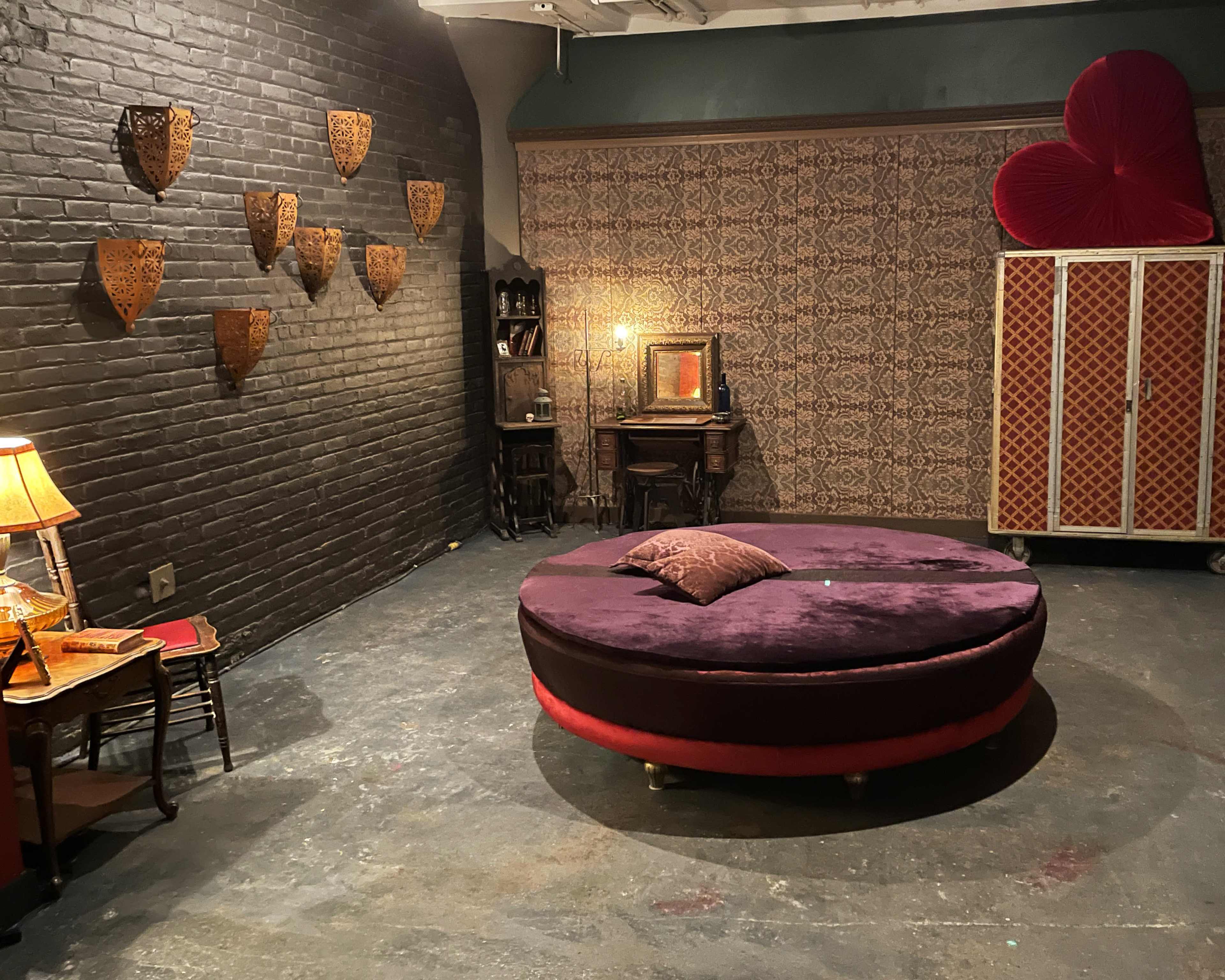 The image depicts a cozy, dimly lit room featuring a round purple velvet sofa, decorative wall art, and a vintage lamp beside a small table.