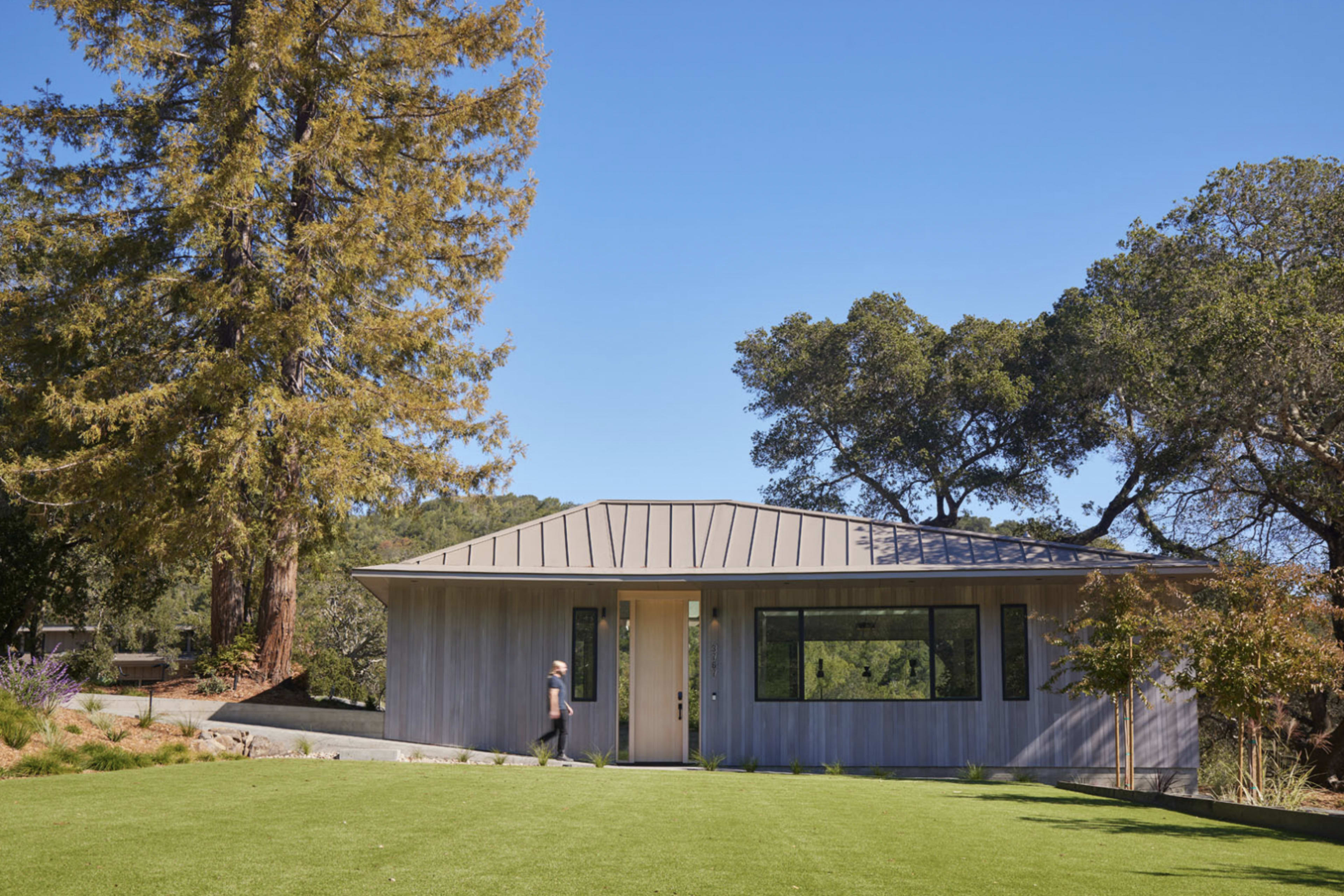 A person walks towards a modern gray house with a metal roof, surrounded by a lawn and trees under a clear blue sky.