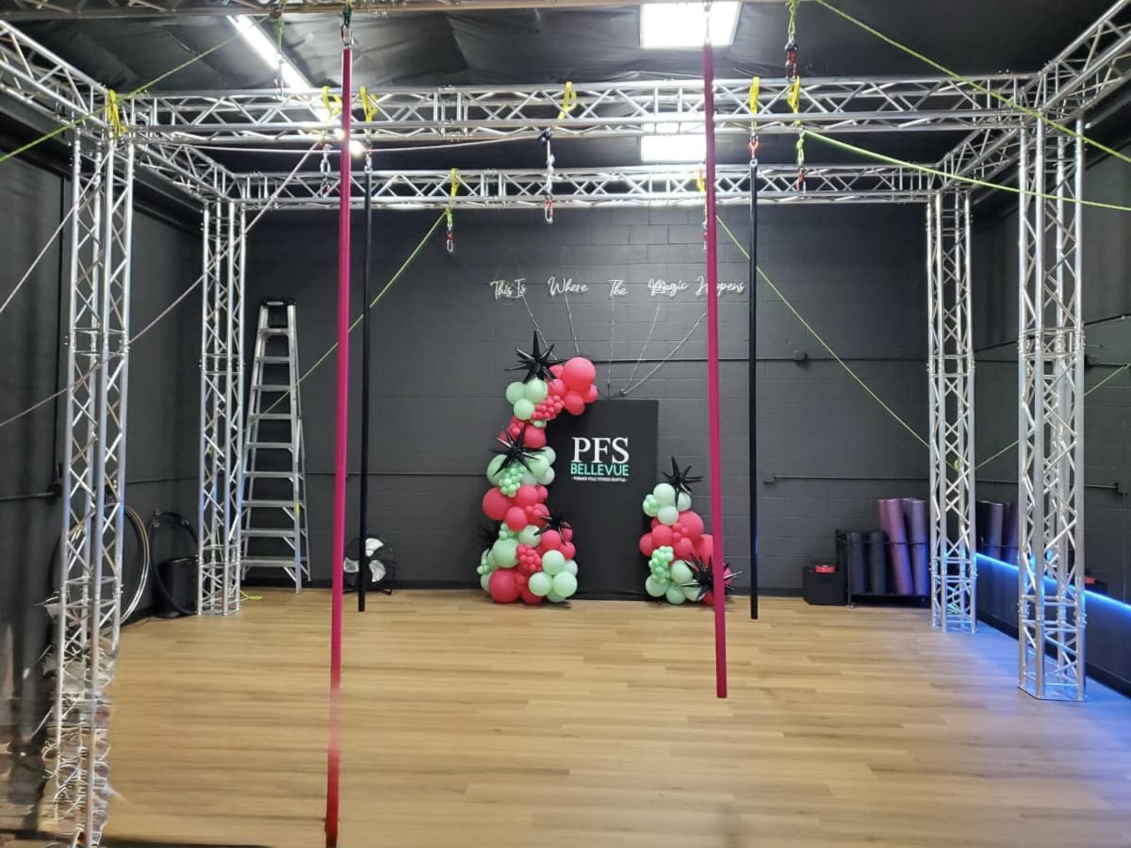 The image shows an indoor fitness space featuring a metal frame structure with hanging apparatus, colorful balloon decorations, and a wooden floor.