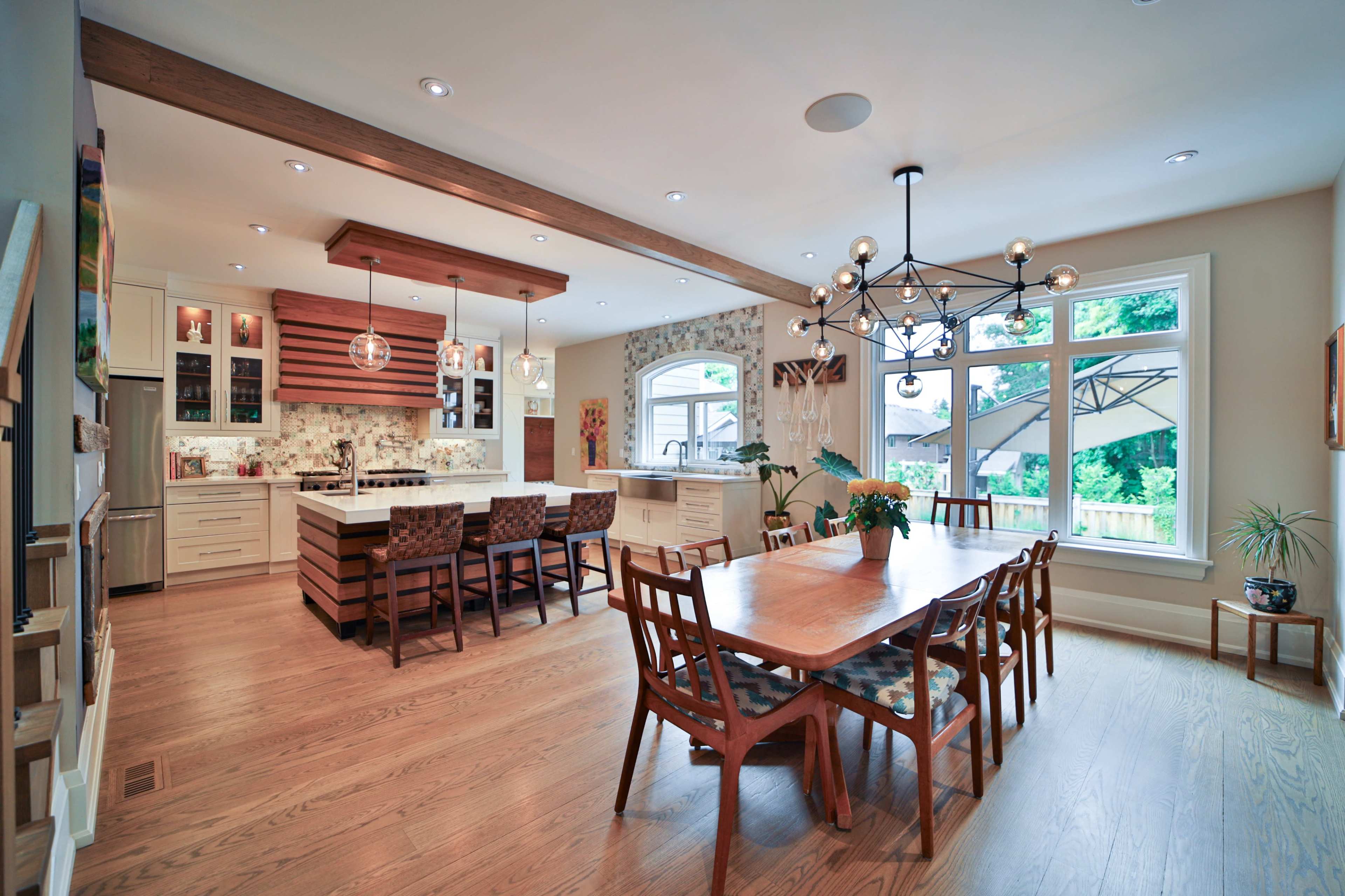A modern kitchen and dining area features a large wooden table surrounded by chairs, a central island with pendant lights, and a view of the outdoors through large windows.