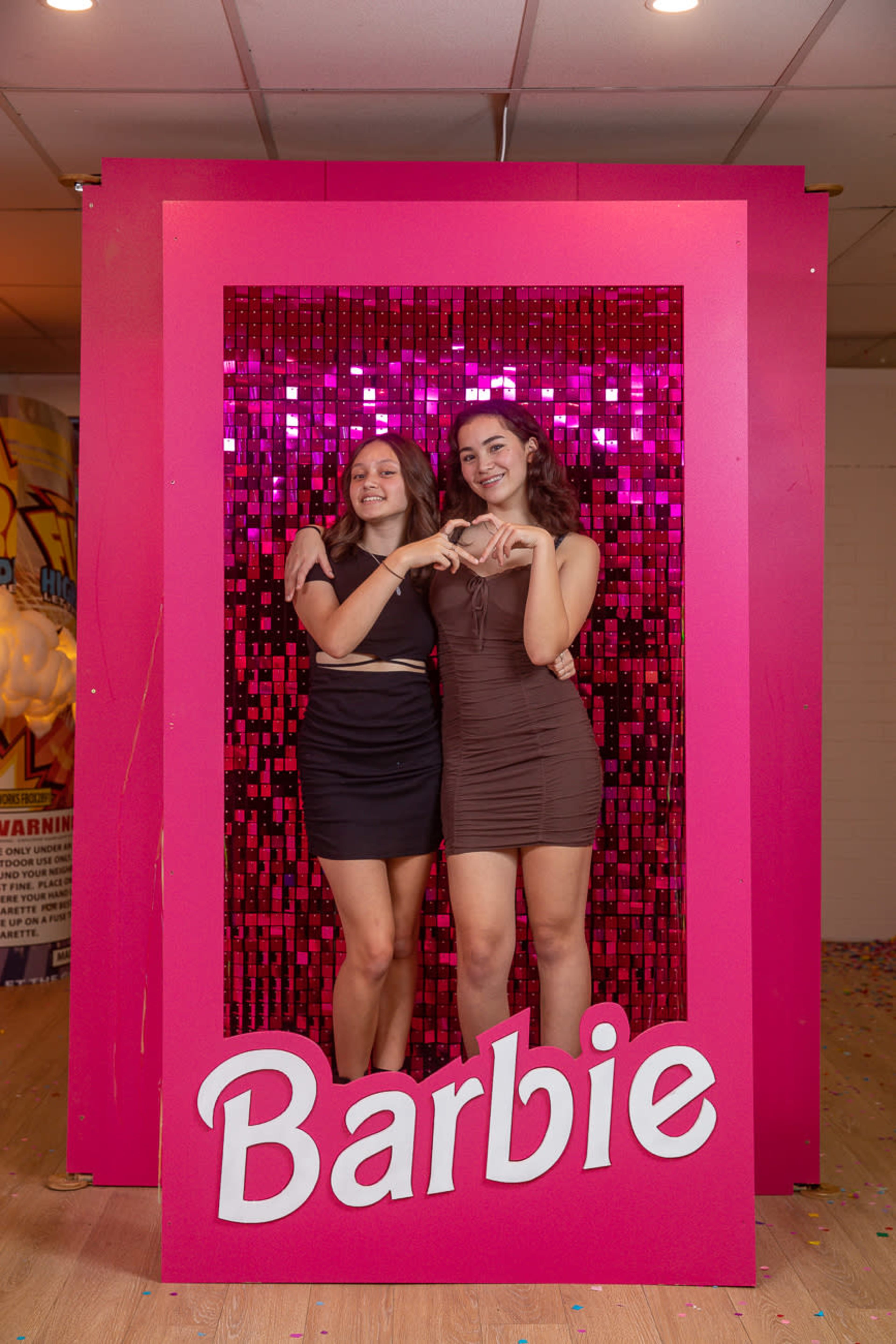 Two young women stand together in a brightly decorated frame with a pink background and "Barbie" written in white at the bottom, creating a playful photo setting.