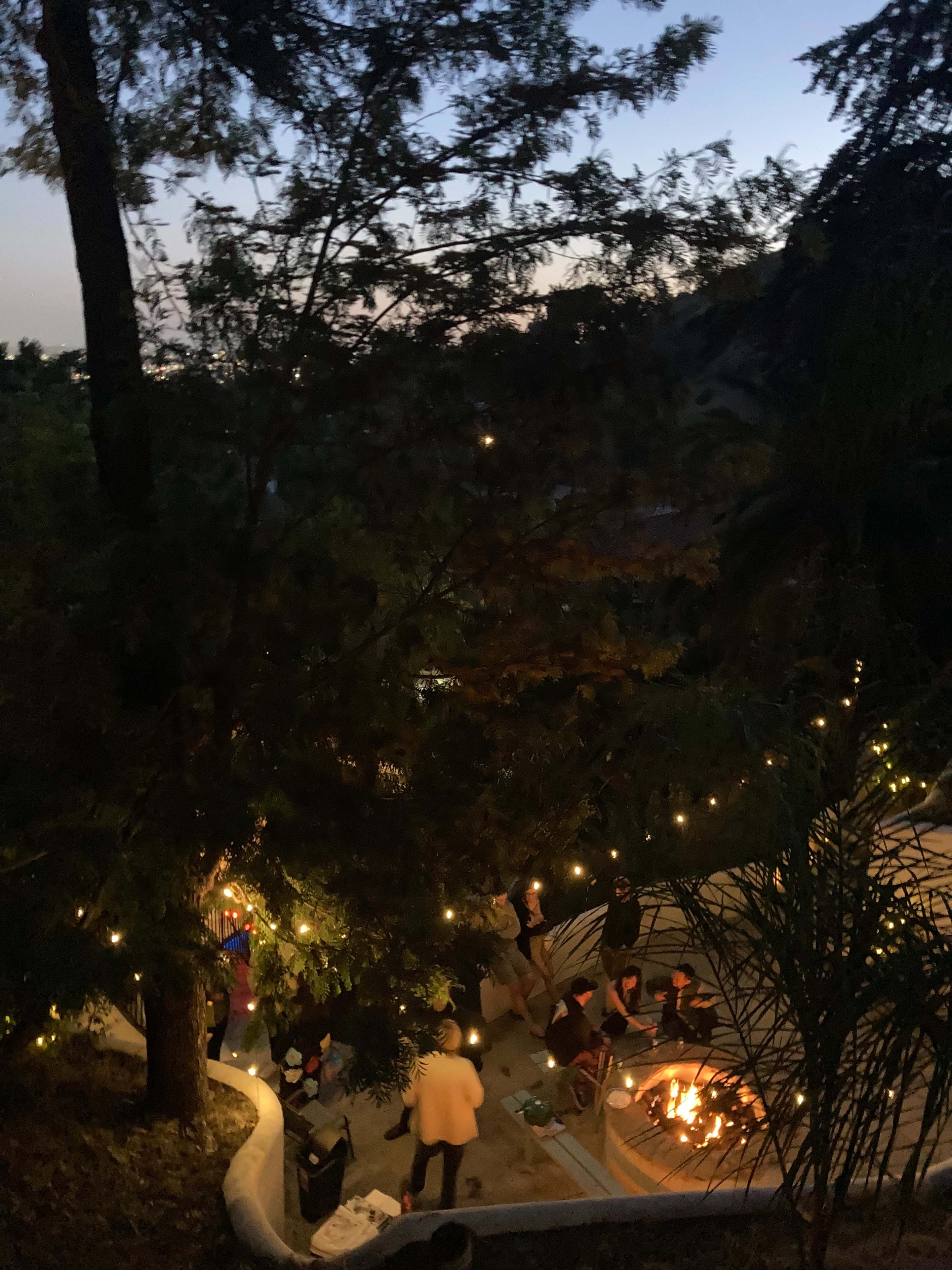 A group of people is gathered around a fire pit surrounded by string lights in a wooded outdoor setting during twilight.