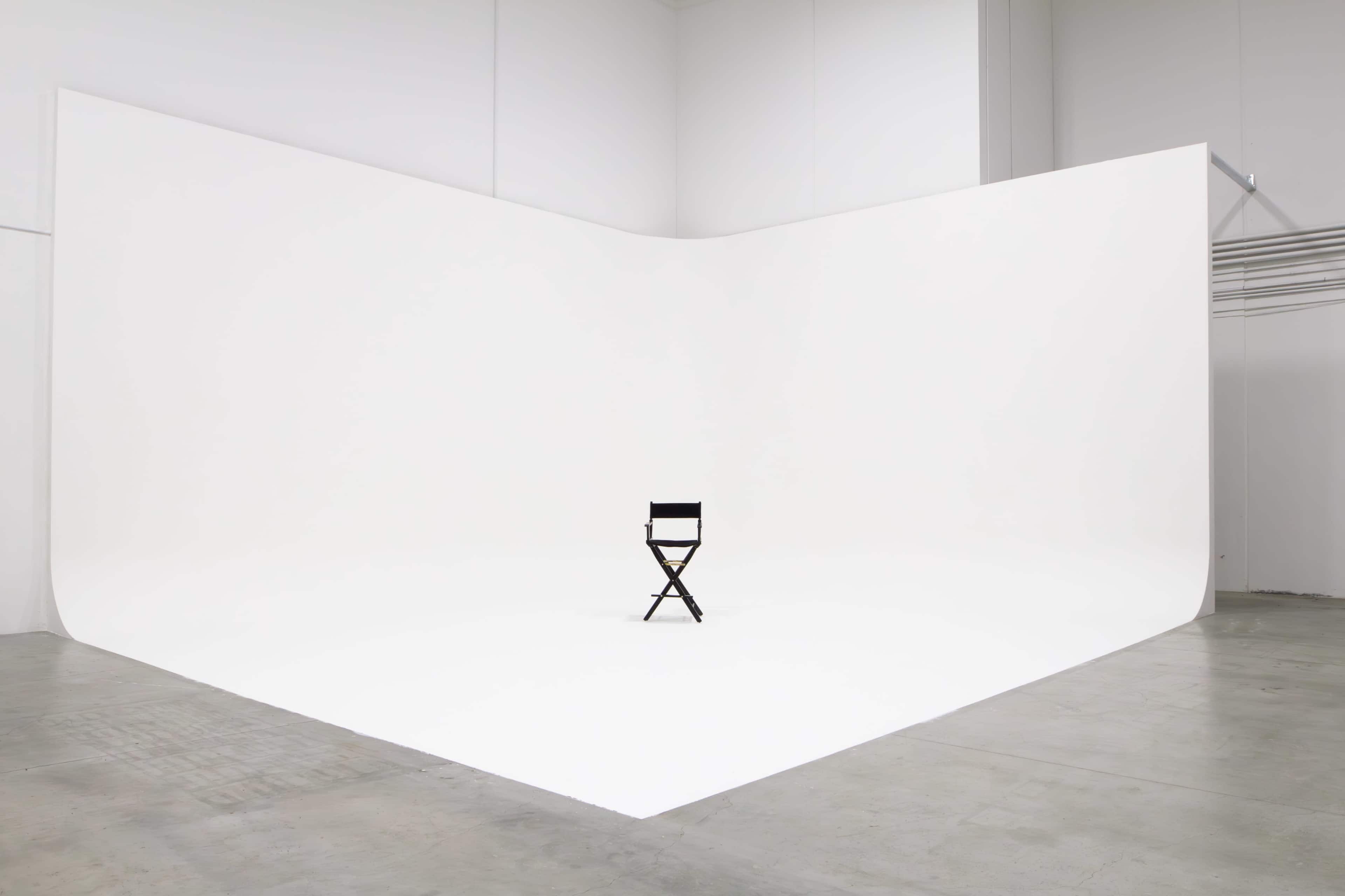 A black director's chair is positioned in the center of a large, empty white studio with a seamless backdrop.