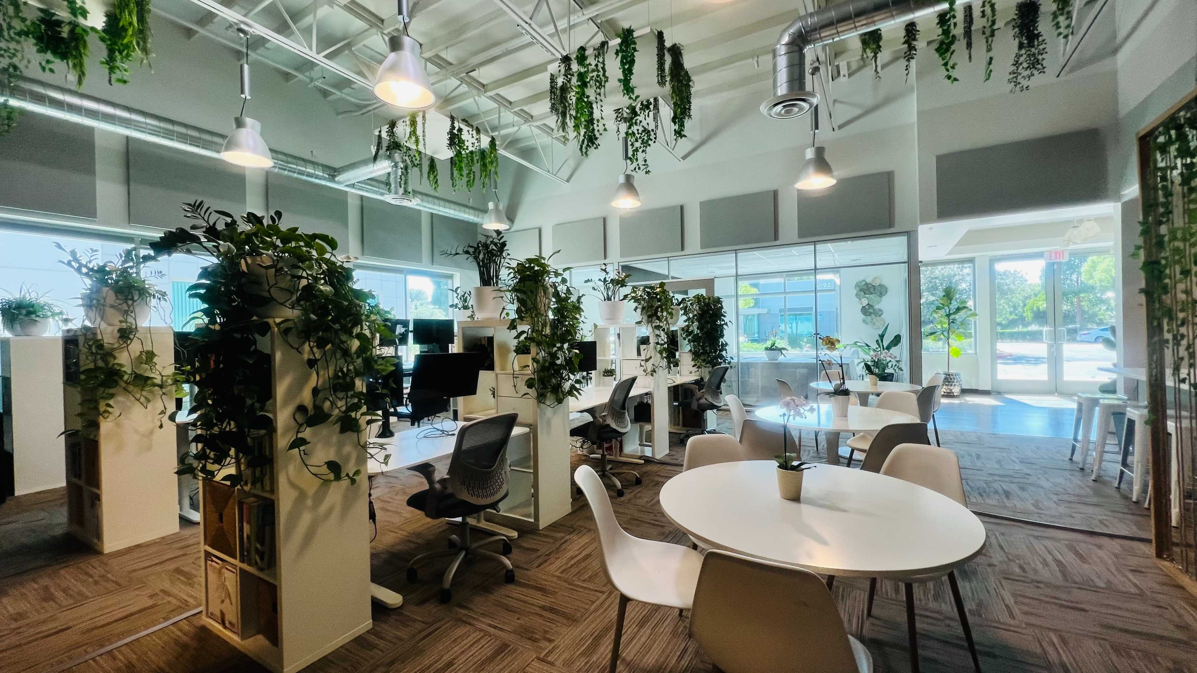 The image shows a modern office space with workstations surrounded by greenery and natural light, featuring round tables and chairs.