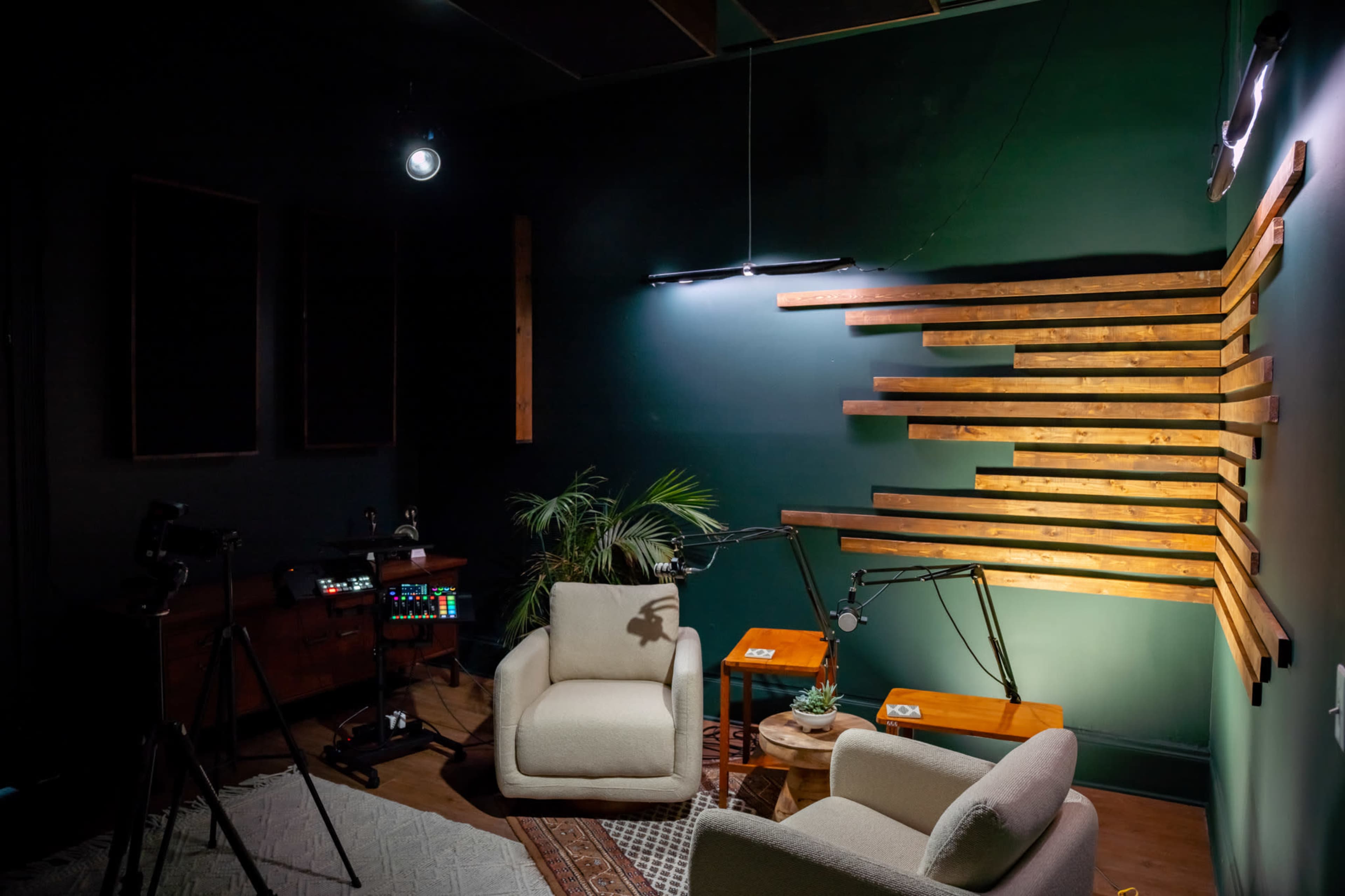 The image shows a dimly lit studio with two light-colored chairs, a wooden accent wall, a small potted plant, and various audio-visual equipment set up on a table.