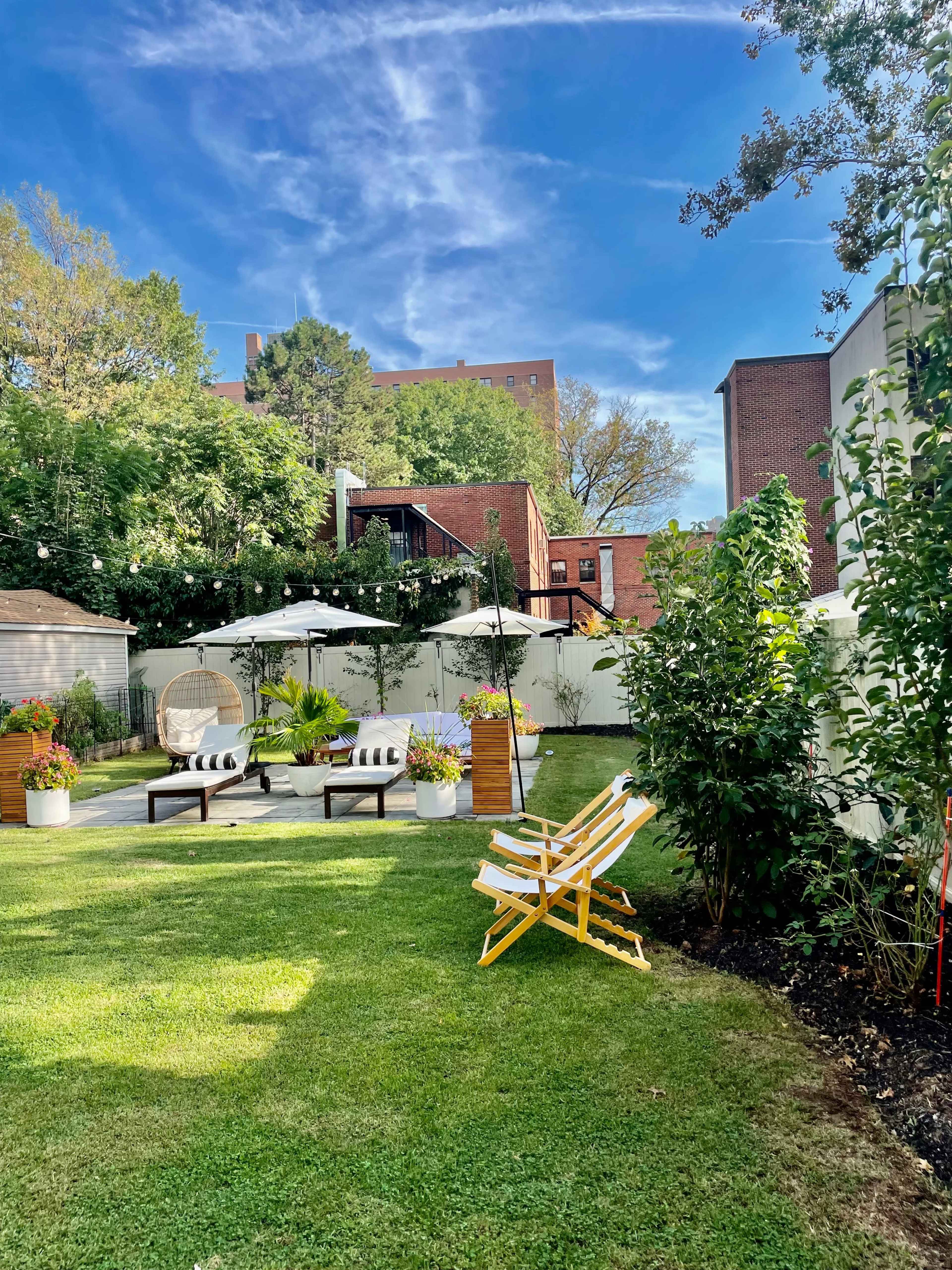 A landscaped backyard with seating areas, umbrellas, and string lights set against a blue sky and greenery.