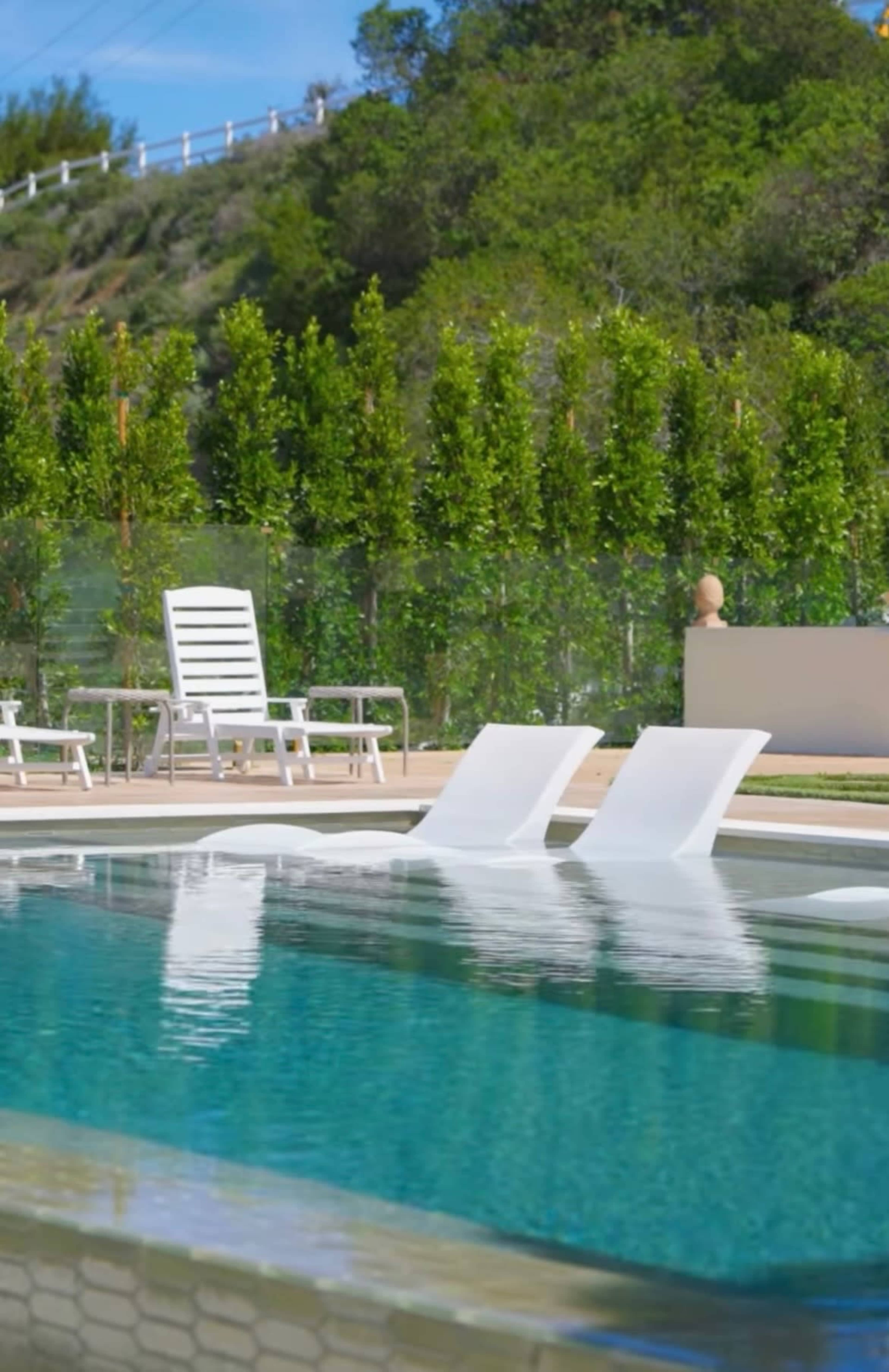 A clear swimming pool with two white lounge chairs sits beside a row of green hedges.
