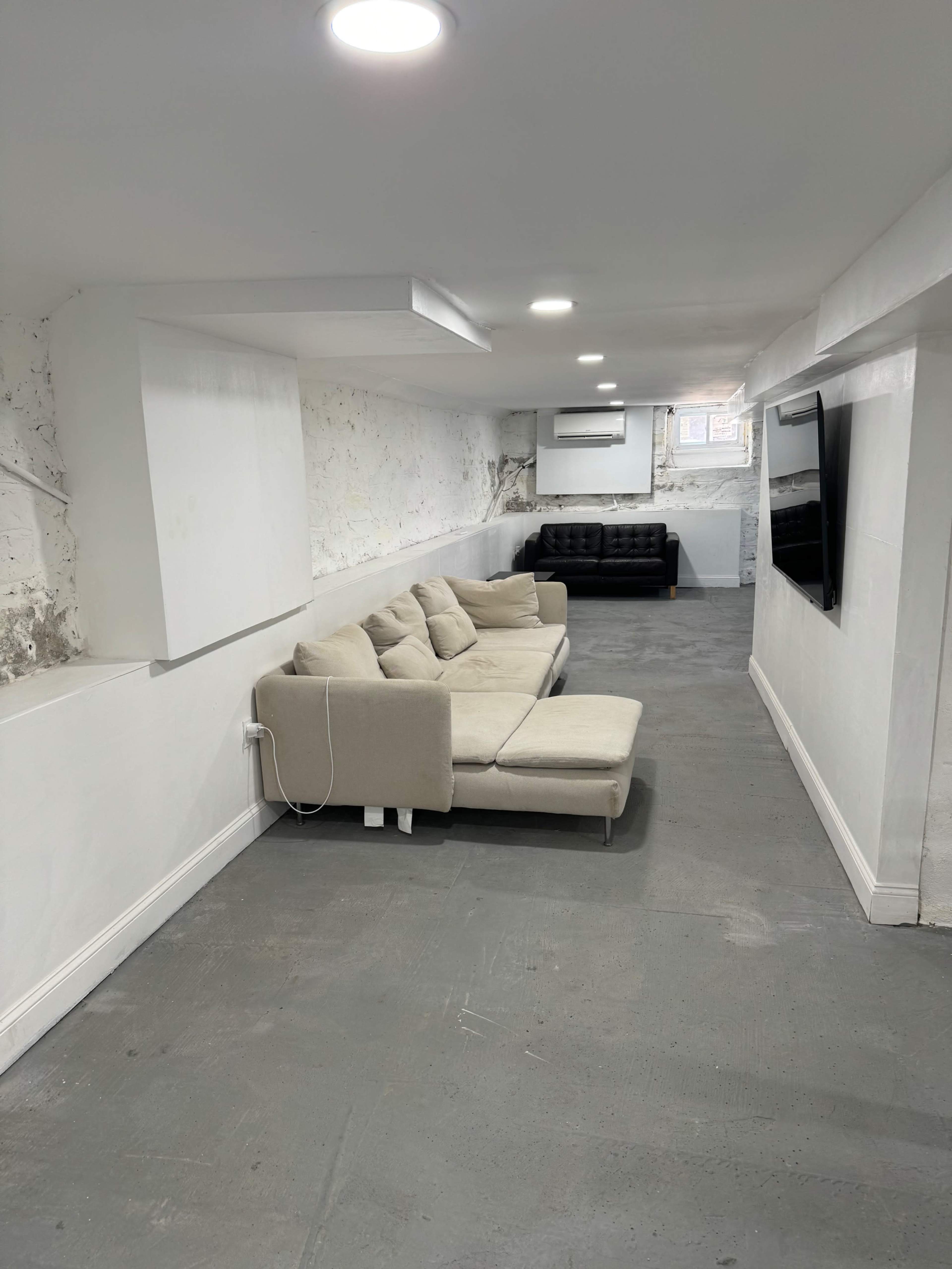 A long, narrow room features a beige sofa and a black leather couch against white walls, with recessed lighting and a television mounted on one side.