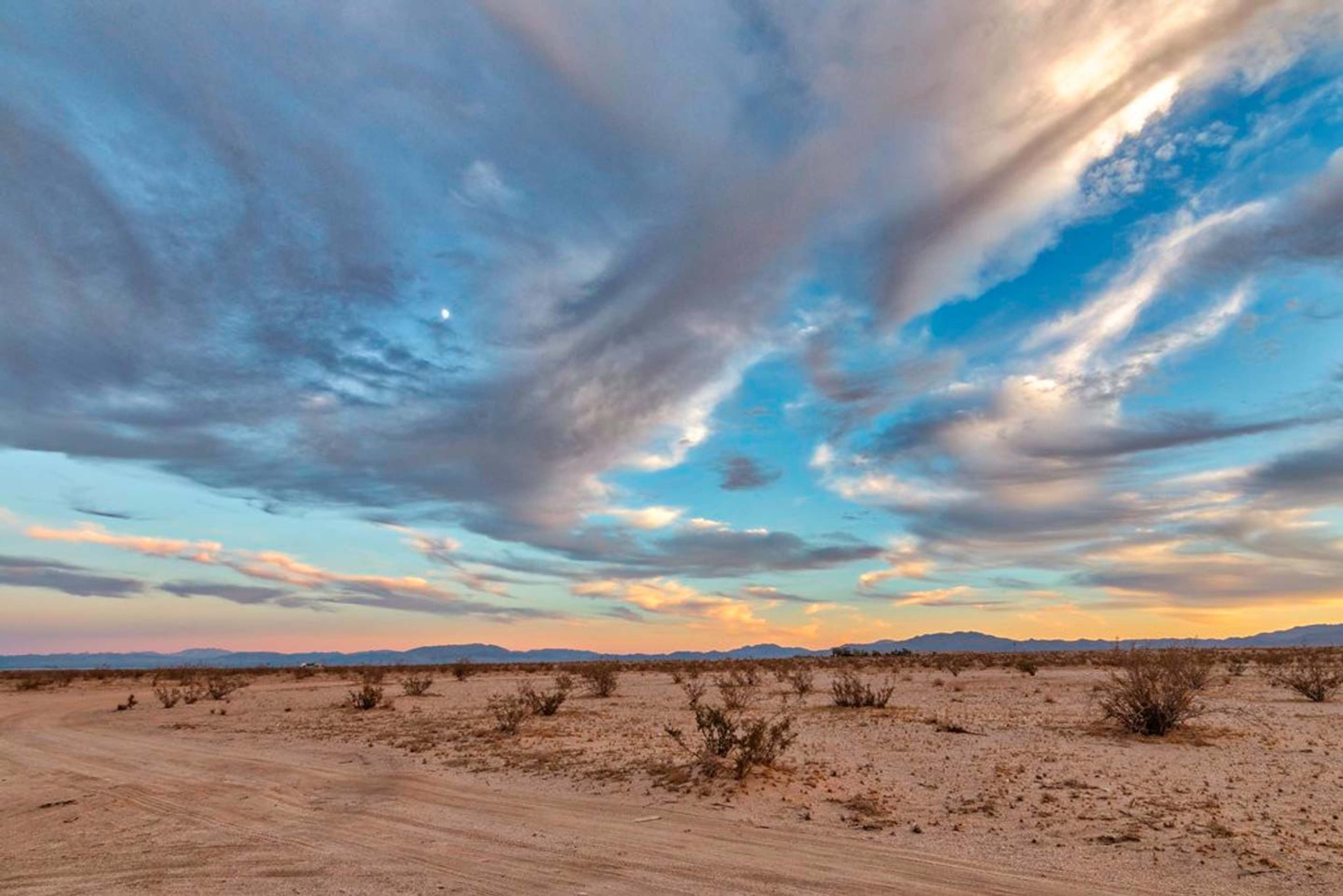 A winding dirt road cuts through a vast desert landscape under a sky filled with swirling clouds.