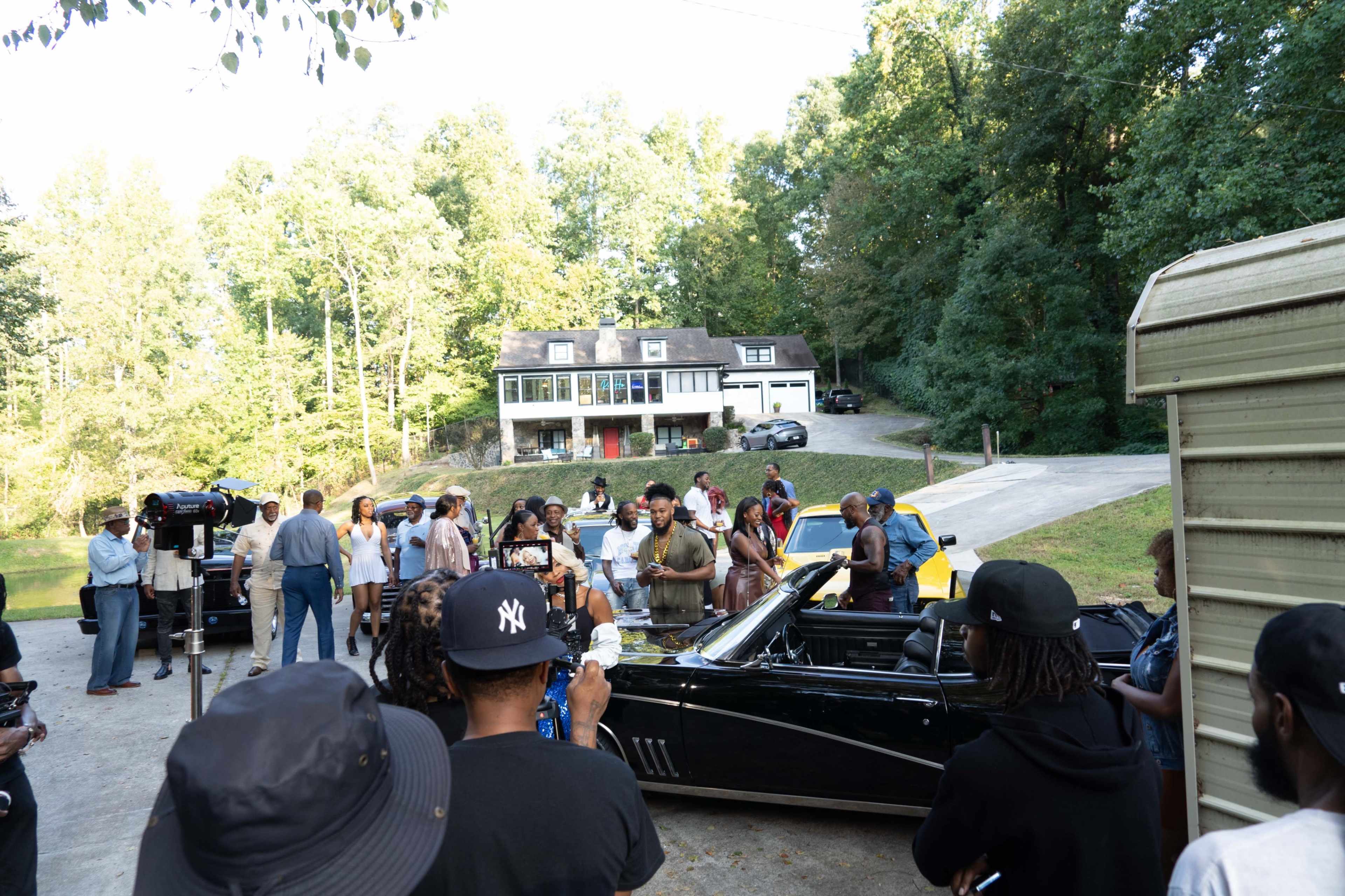A group of people gathers around a black convertible car on a residential street with a large house and several vehicles in the background.
