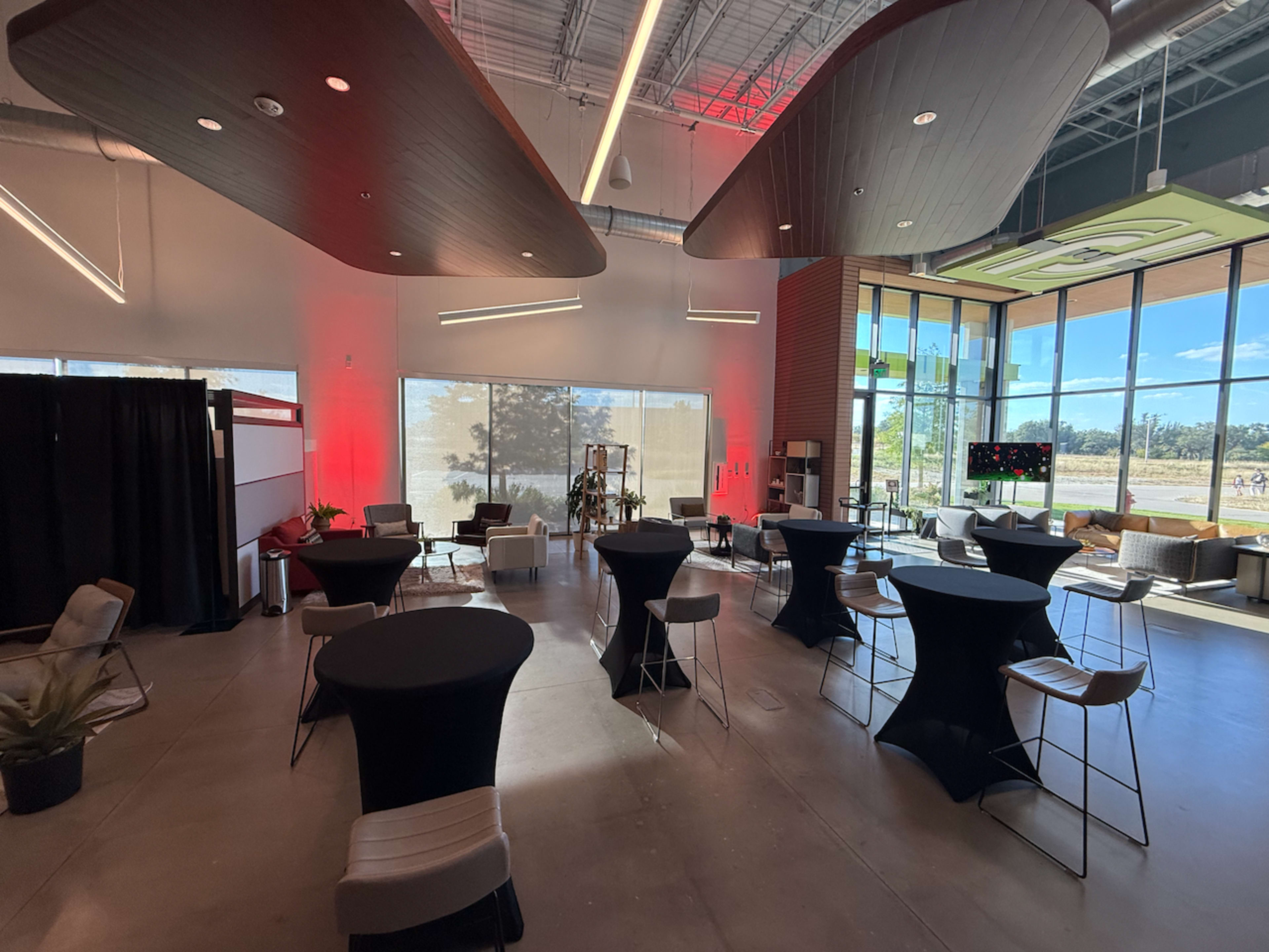 The image depicts a modern event space with tall tables arranged in a partially illuminated room featuring large windows and contemporary architectural design elements.