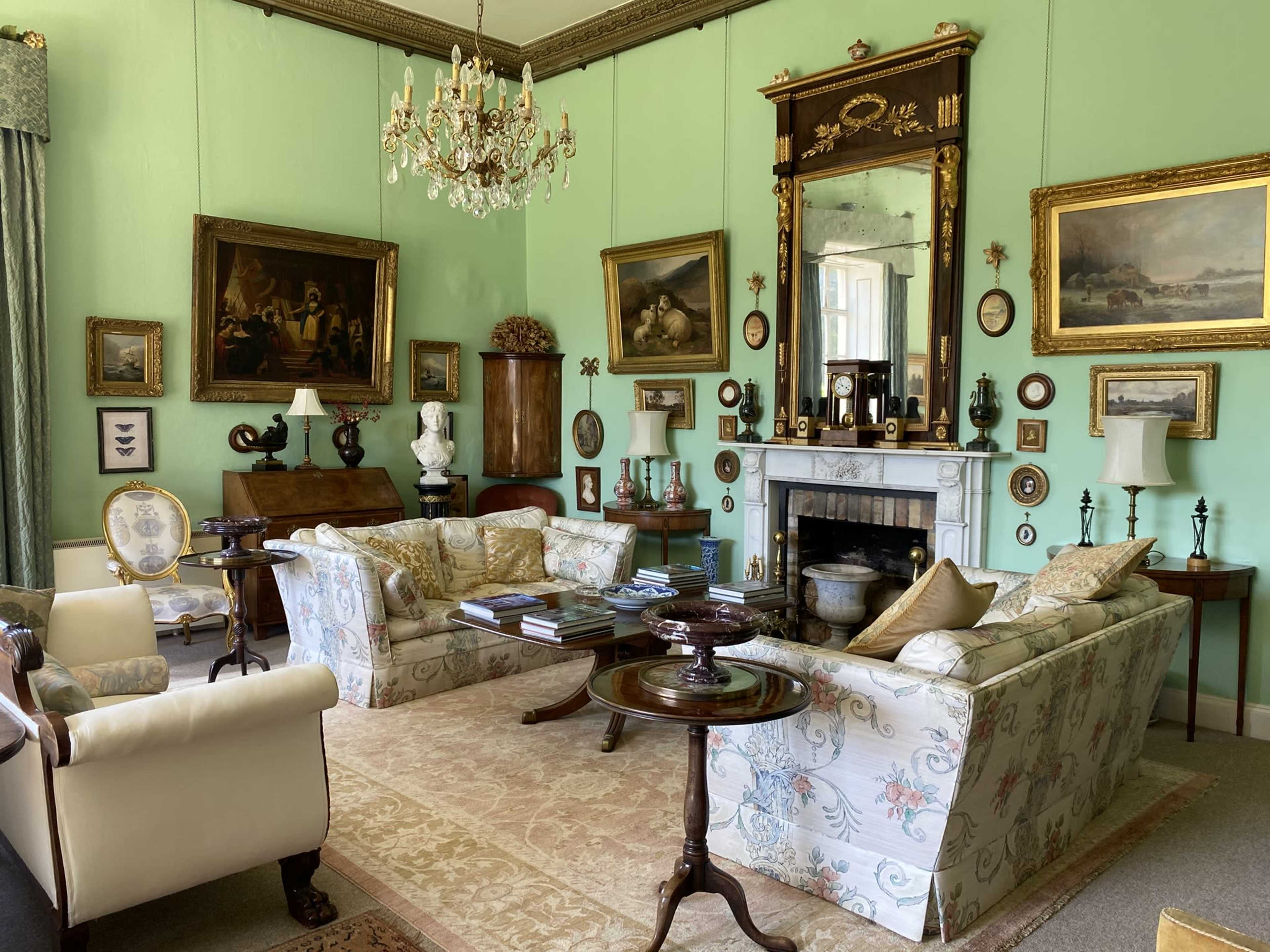 The image shows a well-furnished sitting room with light green walls, two floral-patterned sofas, a chandelier, and various framed artworks displayed on the walls.