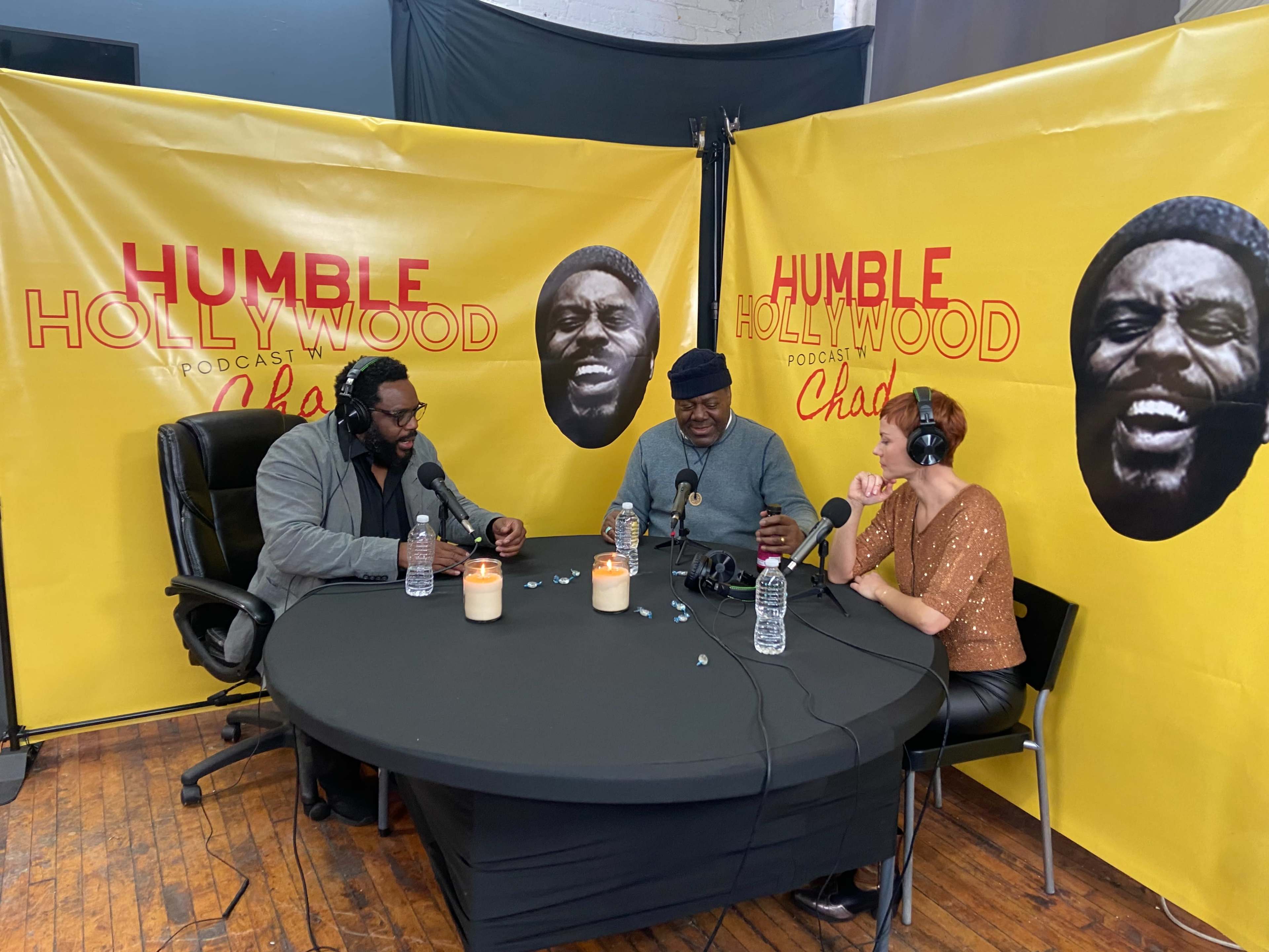 Three people sit at a table during a podcast recording, with yellow walls featuring black-and-white portraits in the background.