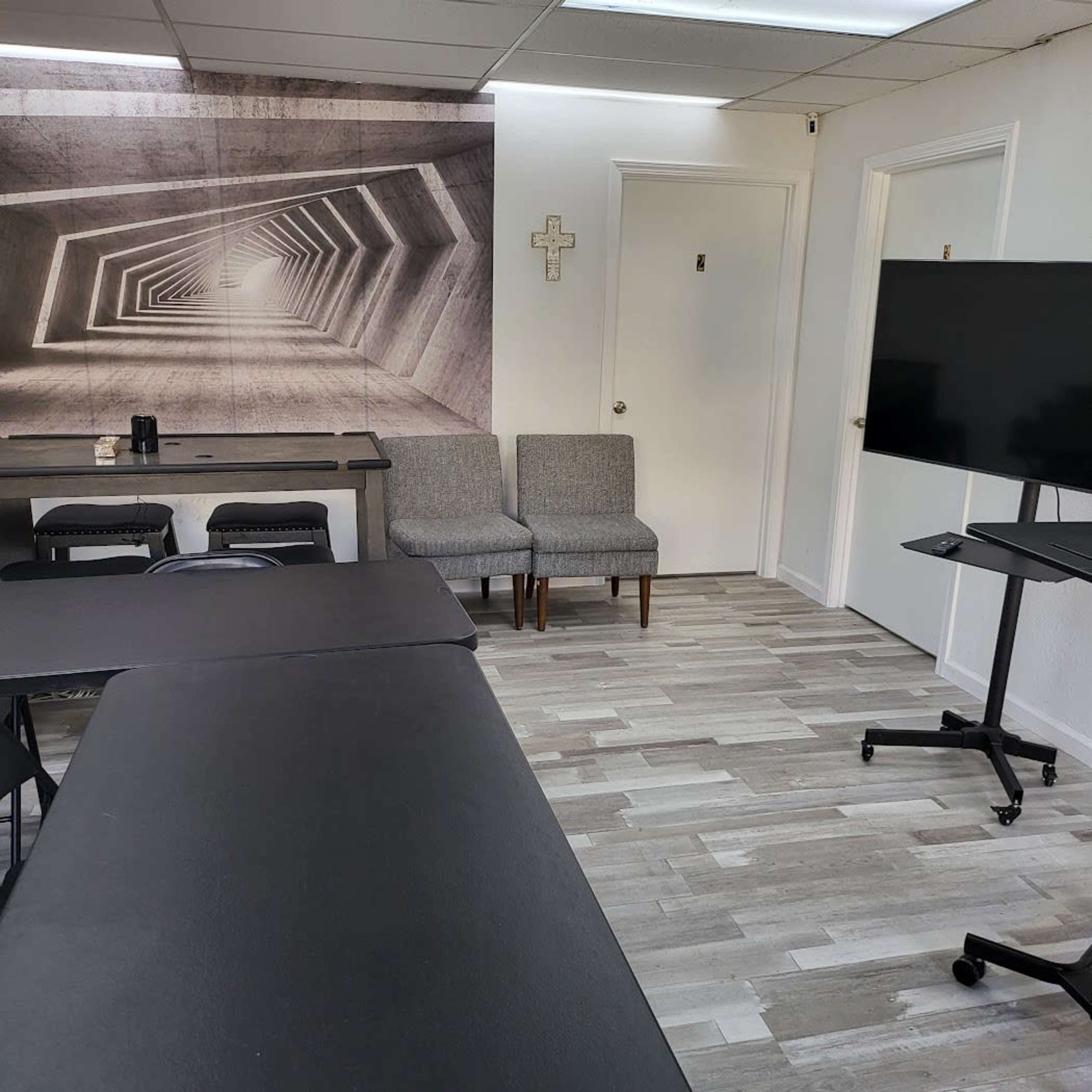 A modern classroom or meeting space with gray walls, a geometric mural, two chairs, and a large display screen on a stand.