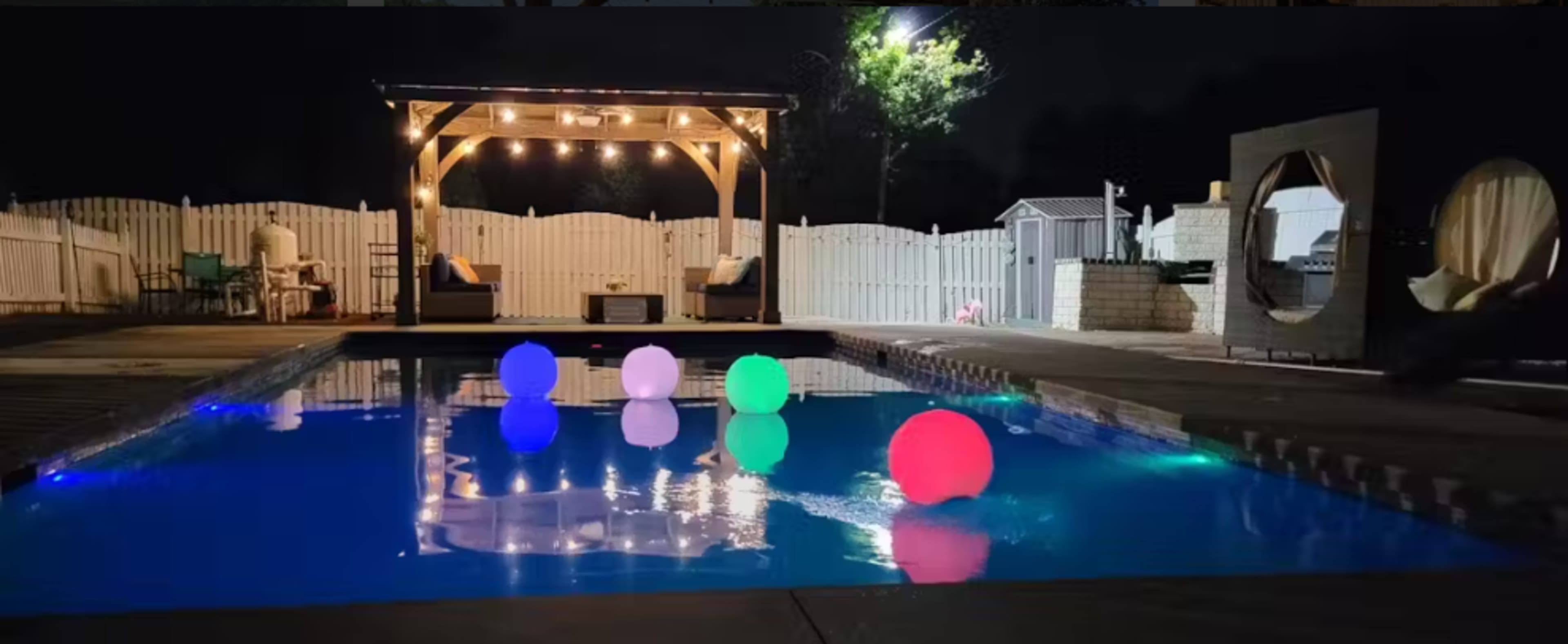 The scene features a swimming pool at night illuminated by colorful floating balls and string lights above a wooden pergola.