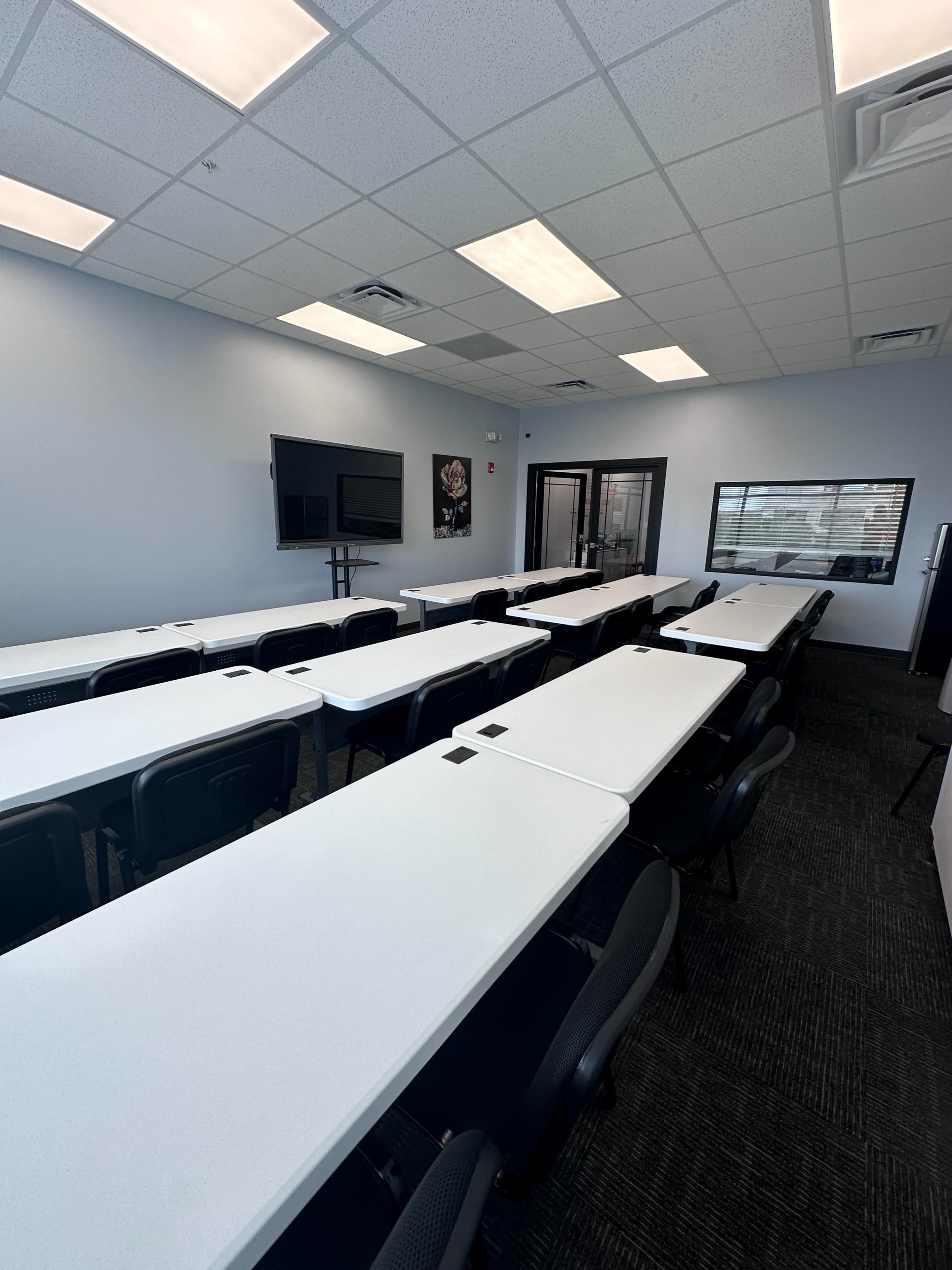 The image shows a modern classroom setup with multiple white tables arranged in rows and a large screen mounted on the wall.