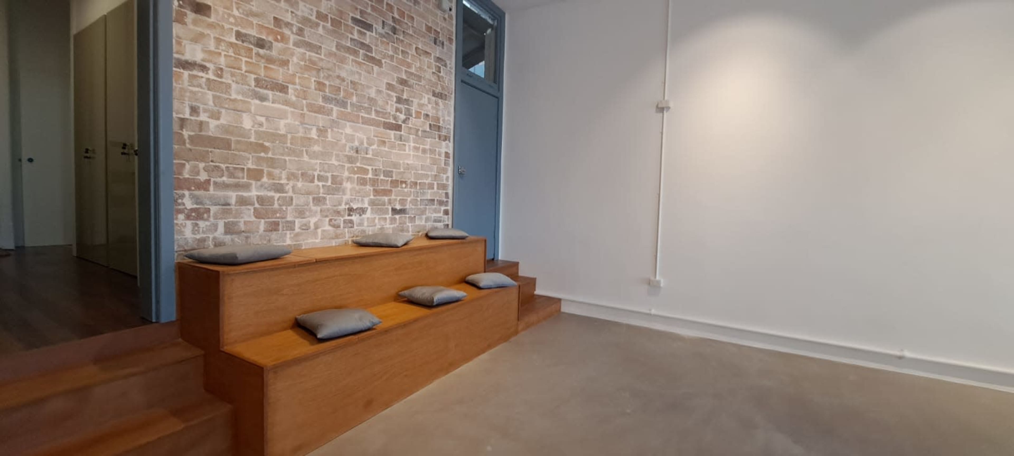 The image shows a minimalistic interior space featuring a wooden bench with cushions against a brick wall and concrete flooring.