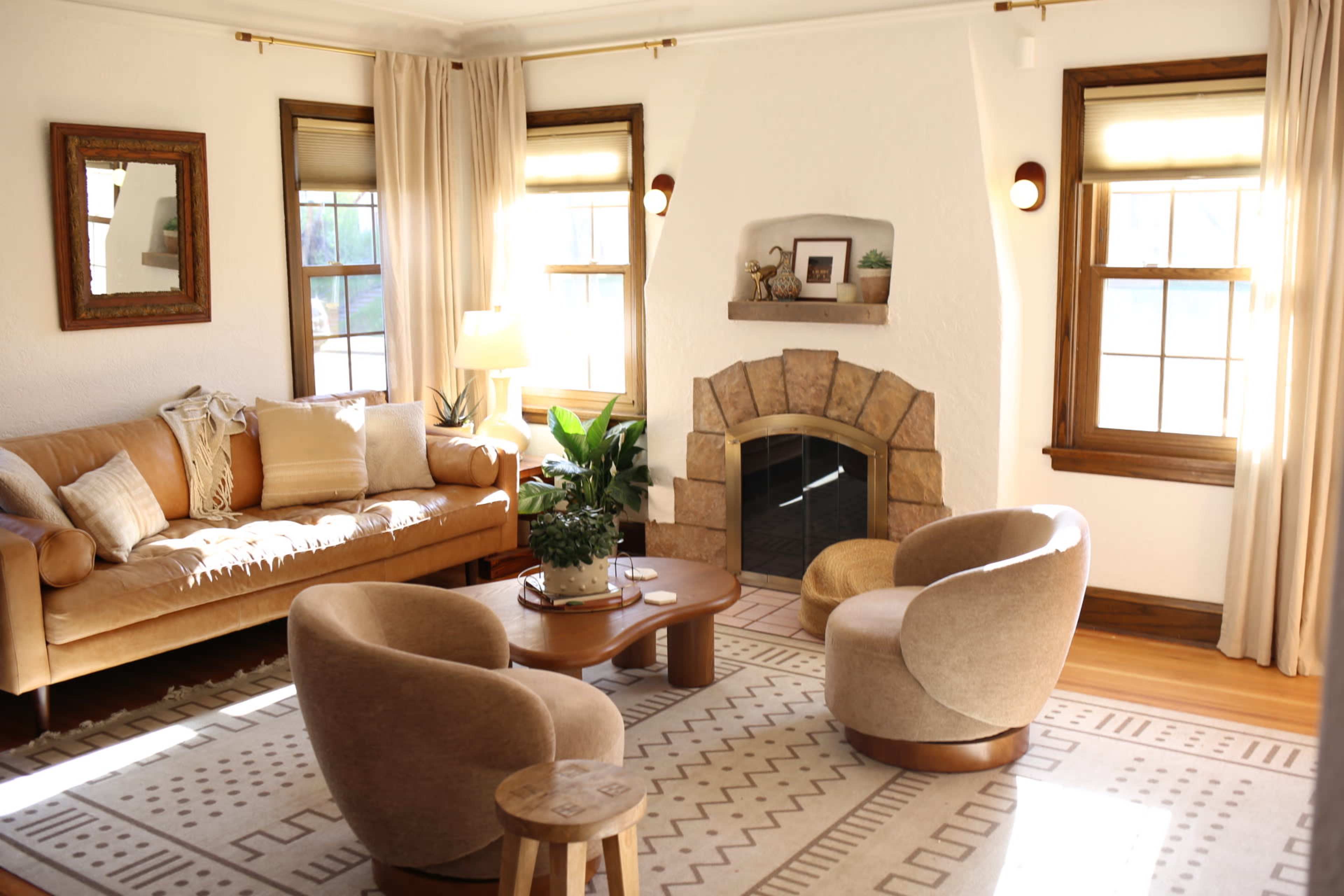 The living room features a brown leather sofa, two circular chairs, a wooden coffee table, and a stone fireplace surrounded by windows with light curtains.