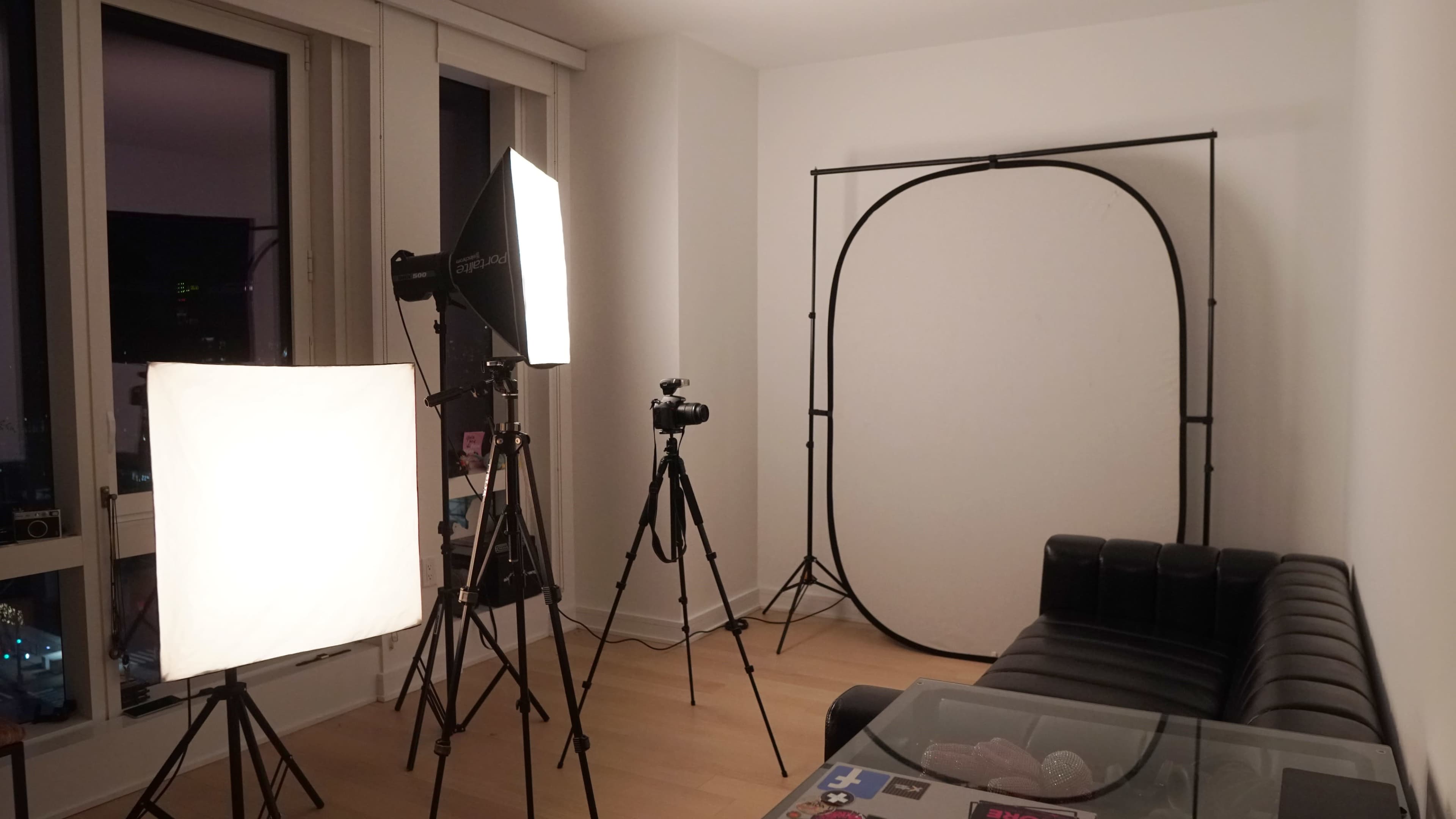 The image shows a photography setup in a bright room, featuring a couch, a backdrop stand with a white backdrop, and multiple lighting equipment on tripods.