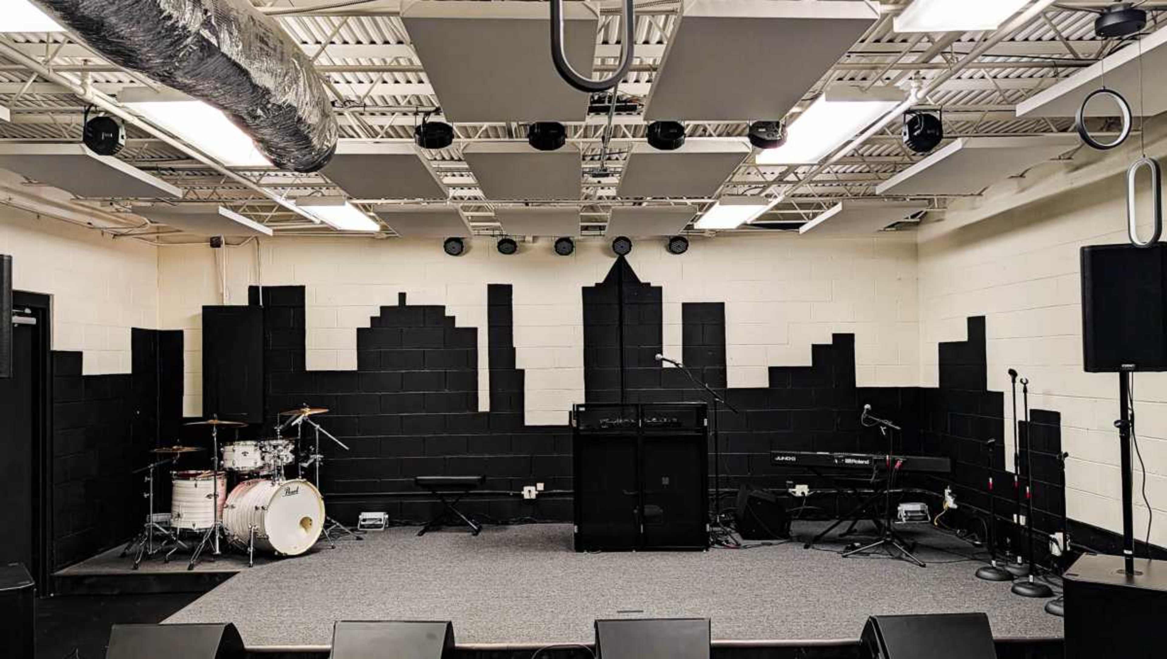 The image shows a music performance space with a black skyline mural on the wall, a drum set, a keyboard, and several microphones set up for a live event.