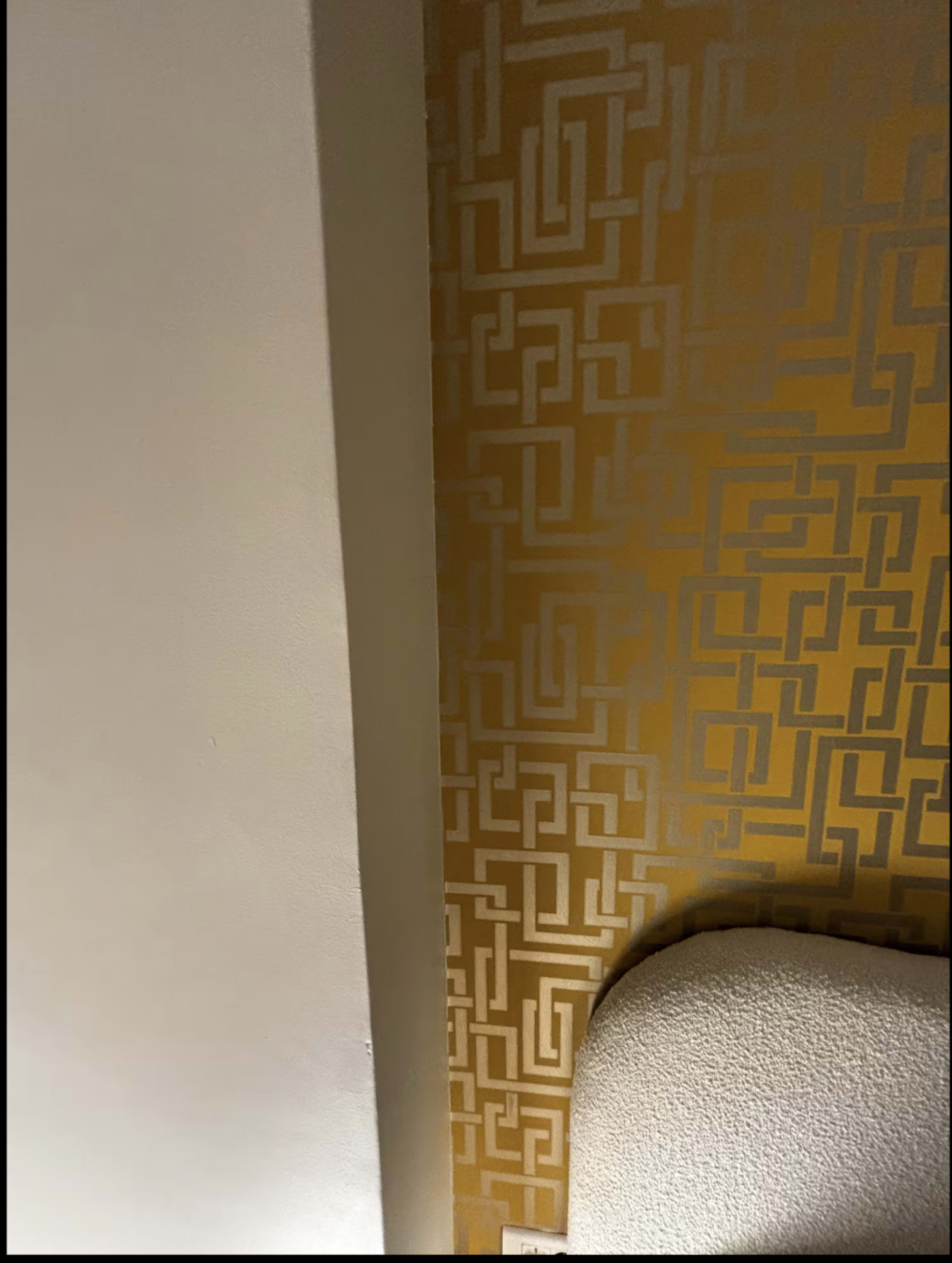 The image shows a corner of a room with a patterned gold and white wallpaper on one wall and a beige upholstered headboard on the adjacent wall.