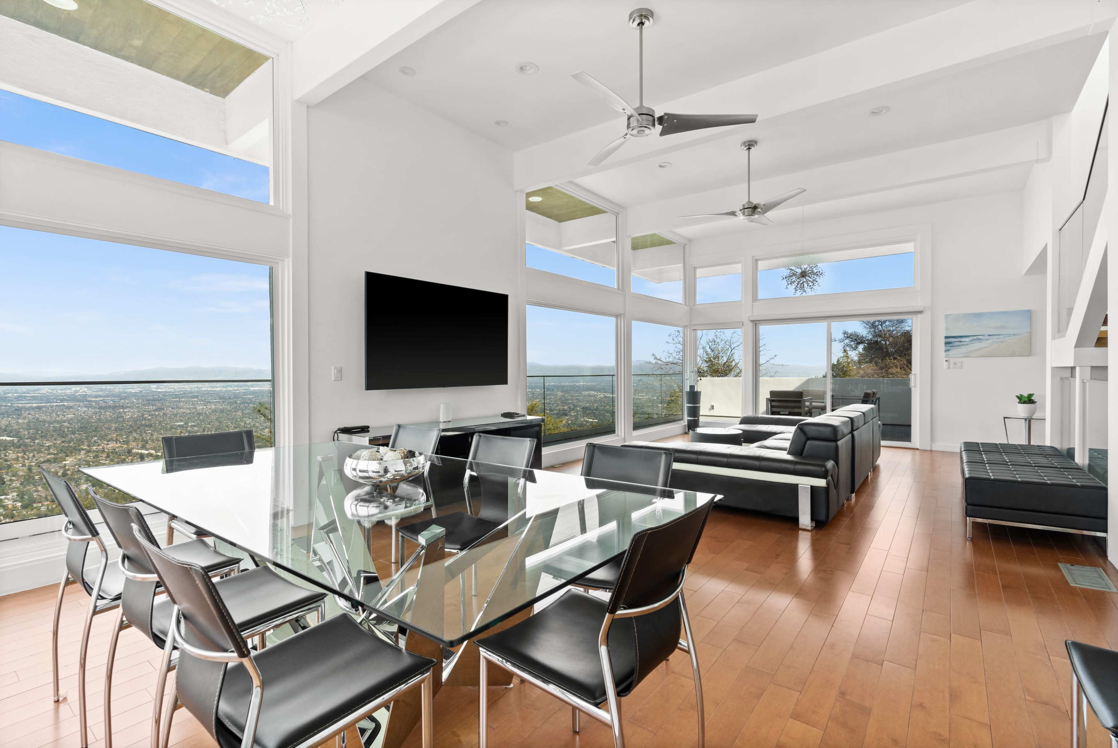 A modern, open-concept living and dining area with large windows overlooking a scenic view, featuring a glass dining table and sleek black furniture.
