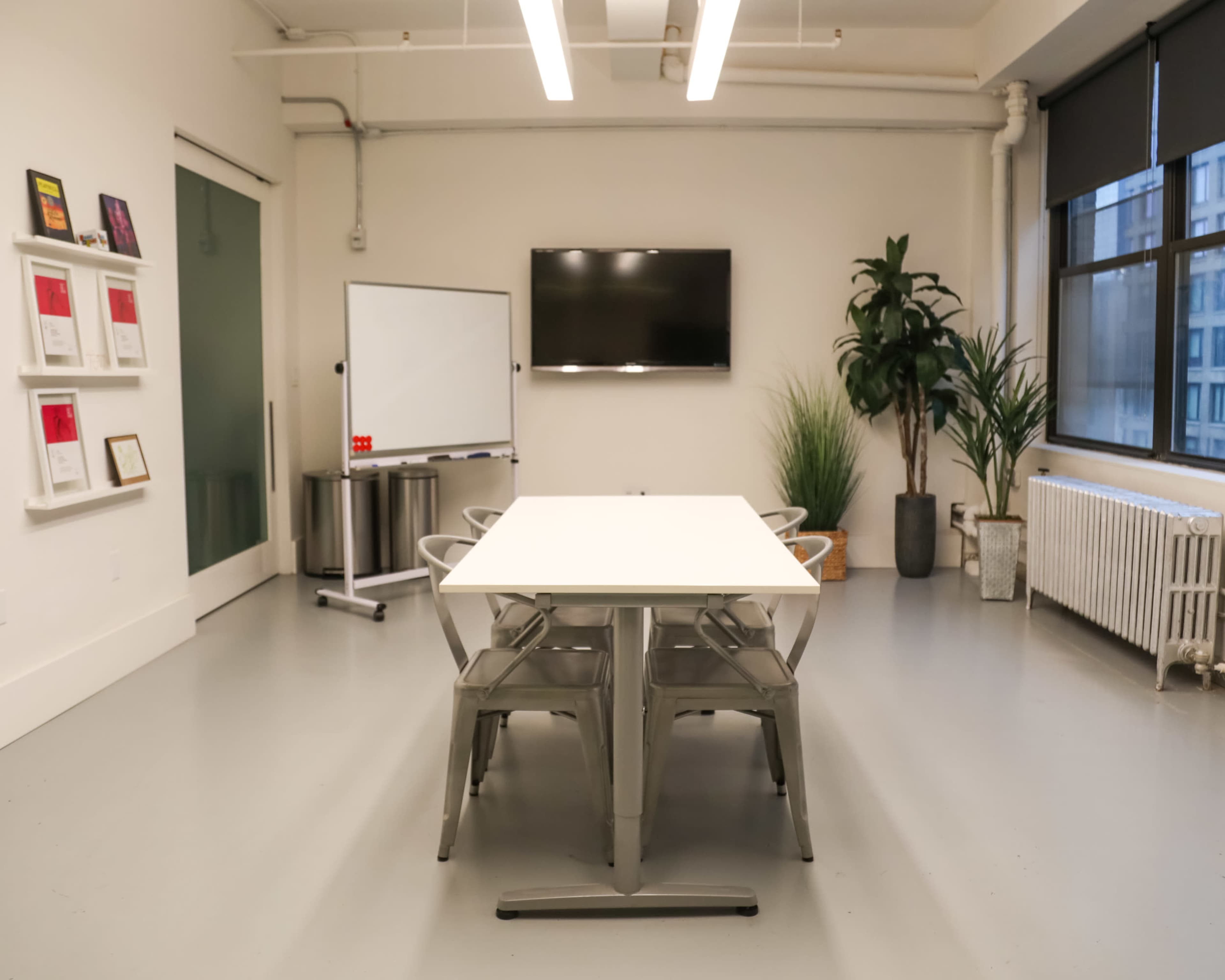 A modern meeting room features a long table surrounded by metal chairs, a whiteboard, a television, and large windows with plants.