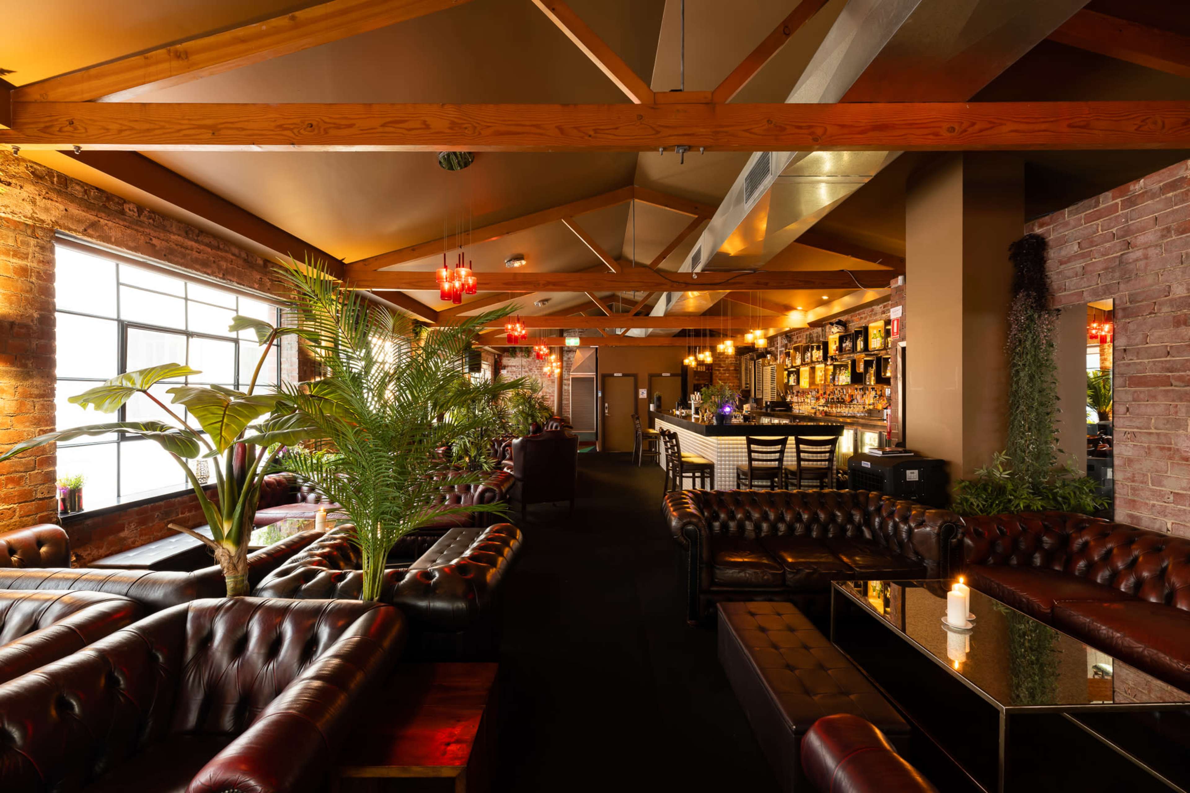A dimly lit bar interior featuring leather couch seating, wooden beams, and a well-stocked bar in the background.