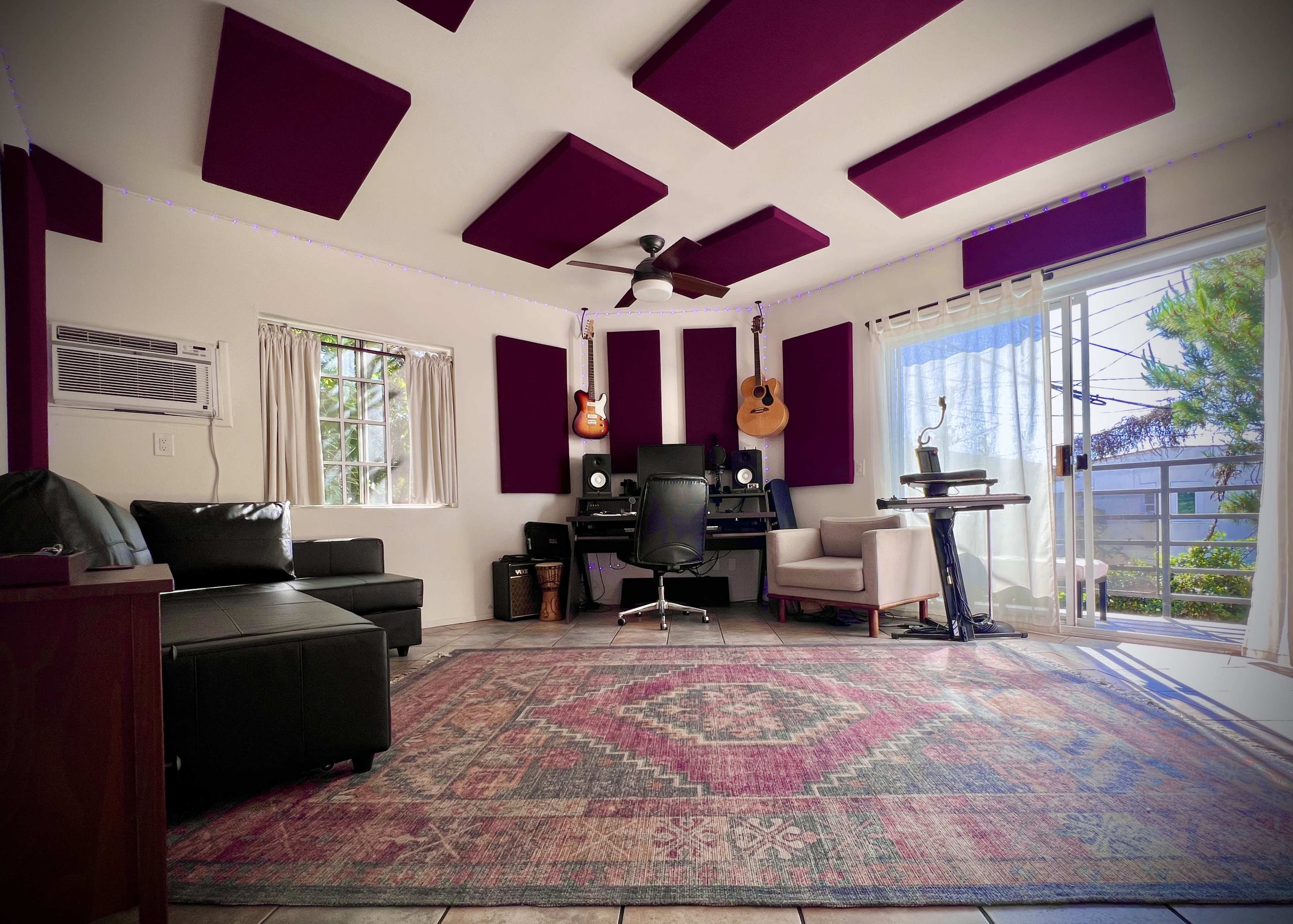 The image shows a home studio with purple acoustic panels, a desk setup with a computer, guitars on the wall, and a cozy seating area.