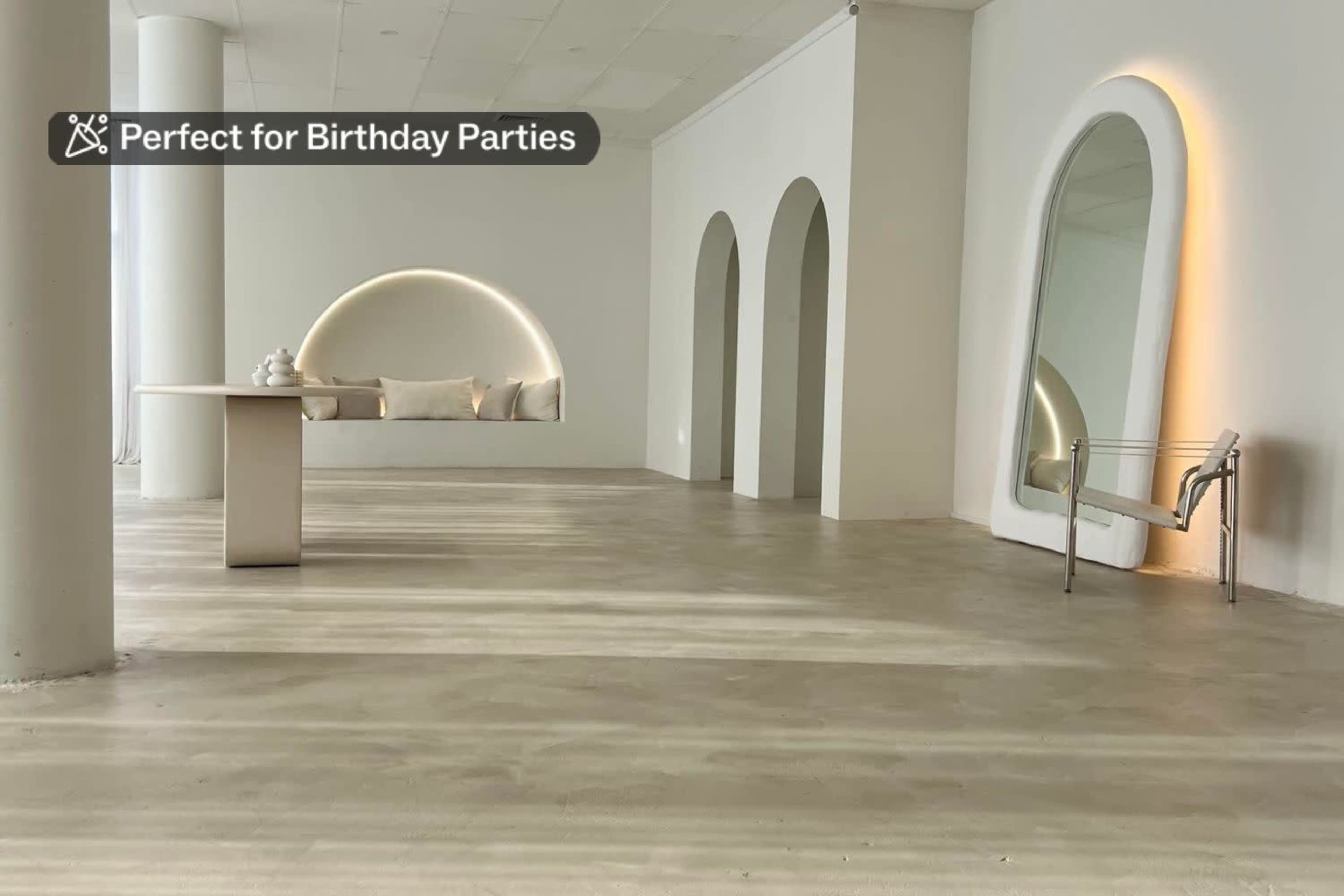 The image shows a minimalistic interior space with a large mirror, a simple table, and archways on the walls, featuring a light-colored floor and soft seating.