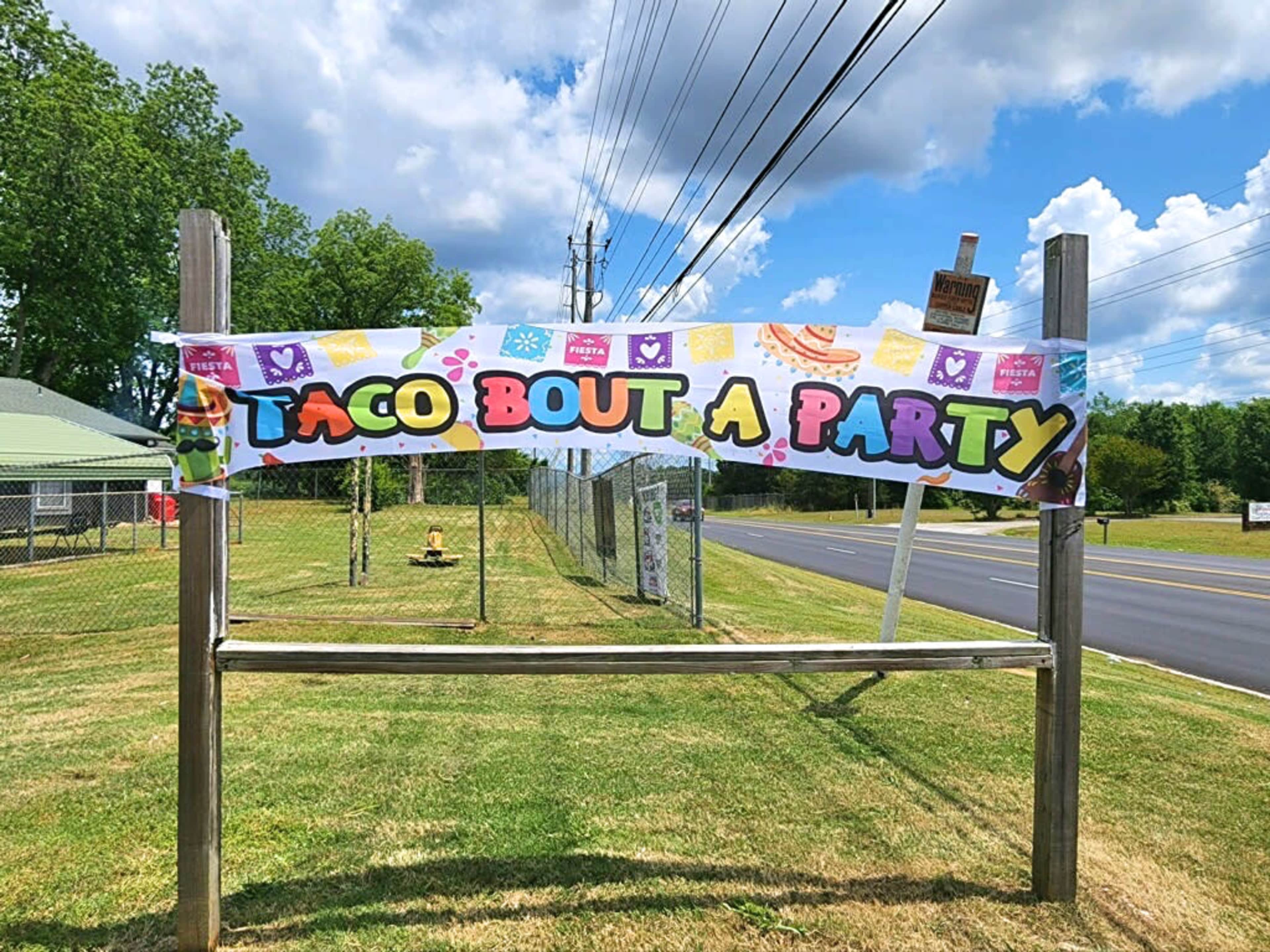 A colorful banner reading "TACO BOUT A PARTY" is displayed on a wooden frame along a grassy area next to a road.