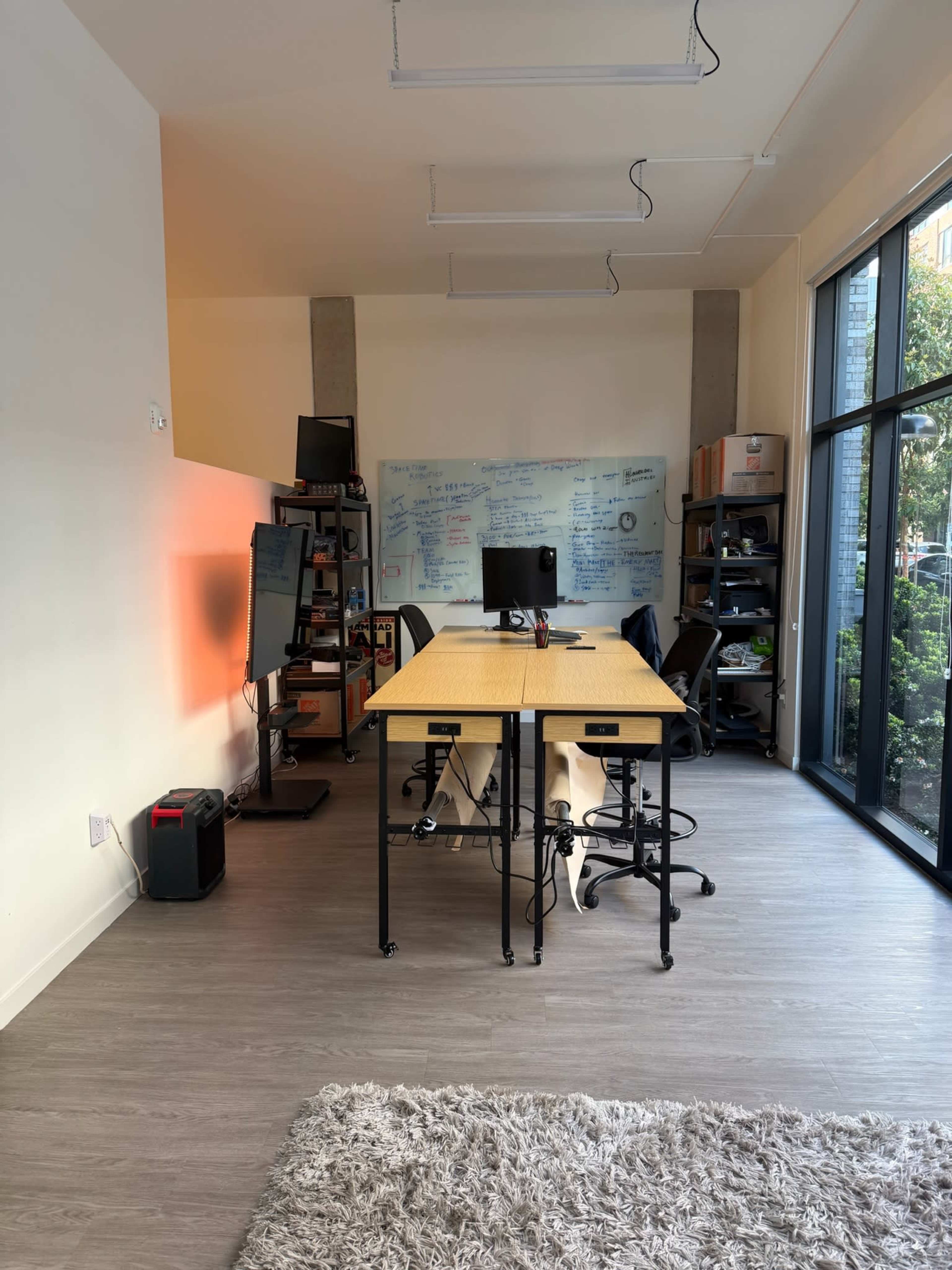 The image depicts a modern office space featuring a large table with four chairs, a black monitor on the table, a whiteboard covered in notes along the back wall, and large windows allowing natural light to enter.
