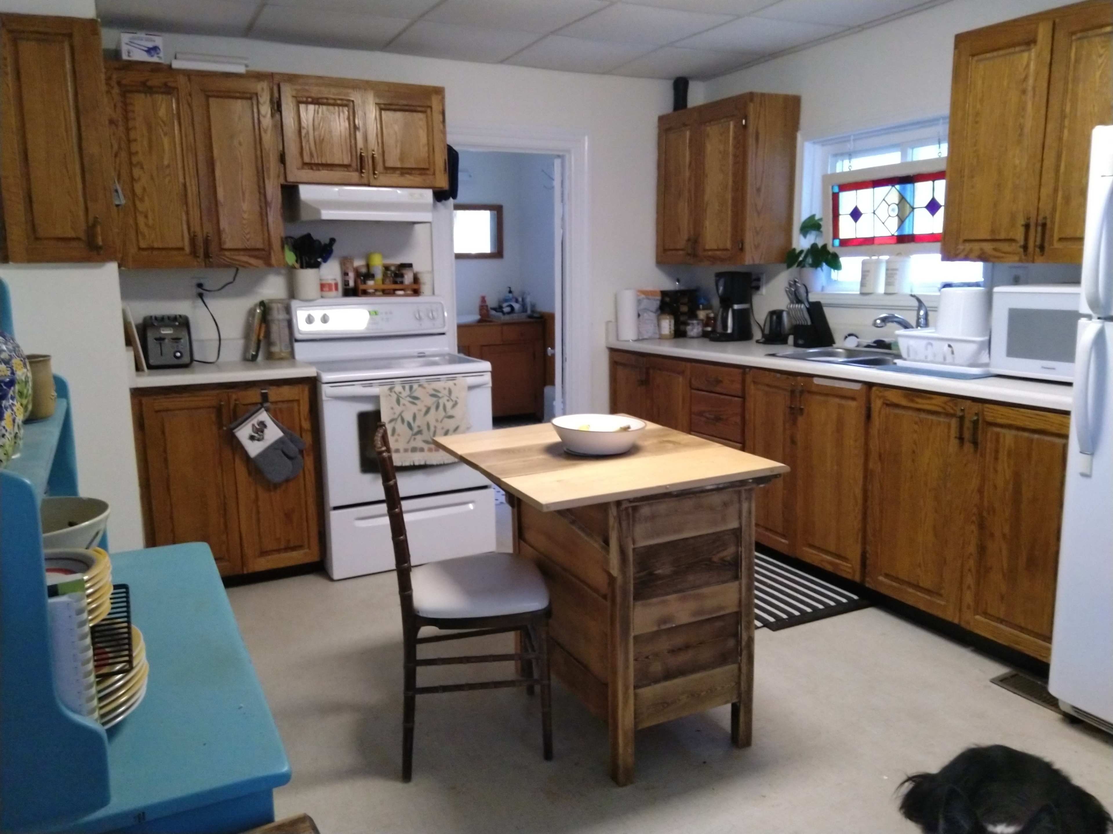 A compact kitchen features wooden cabinets, a small dining table with a bowl, and various appliances along the countertops.