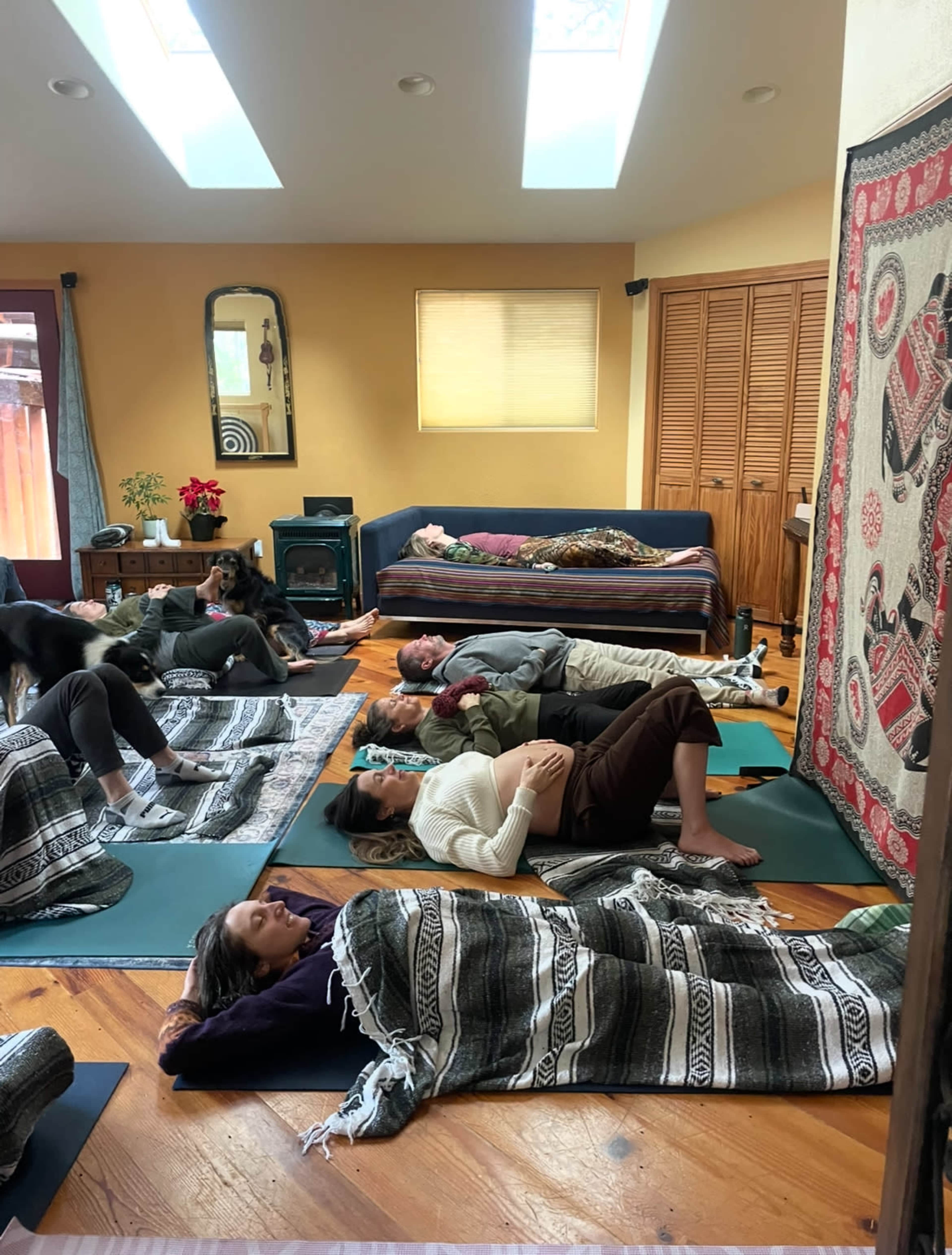 A group of individuals lies on yoga mats, covered with blankets, in a spacious room with natural light and various furnishings.