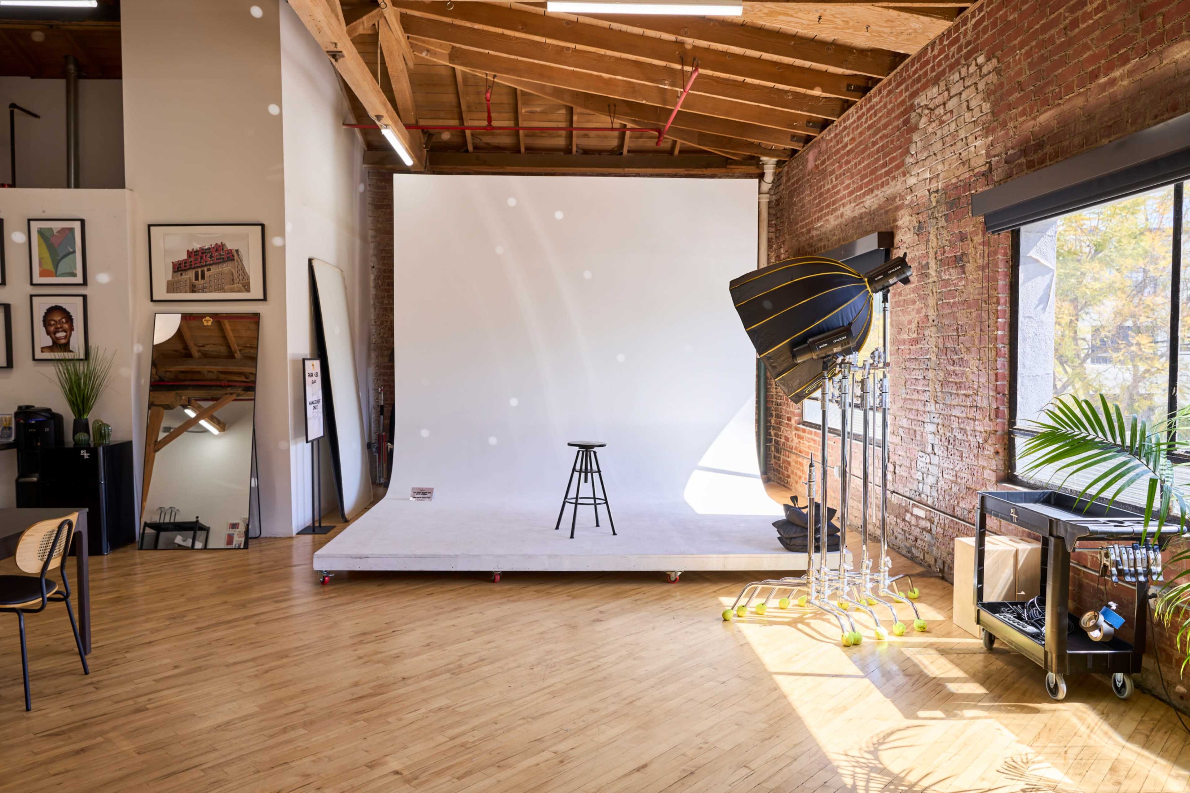 The image shows an empty photography studio with a white backdrop, a stool in the center, and large windows allowing natural light to fill the space.