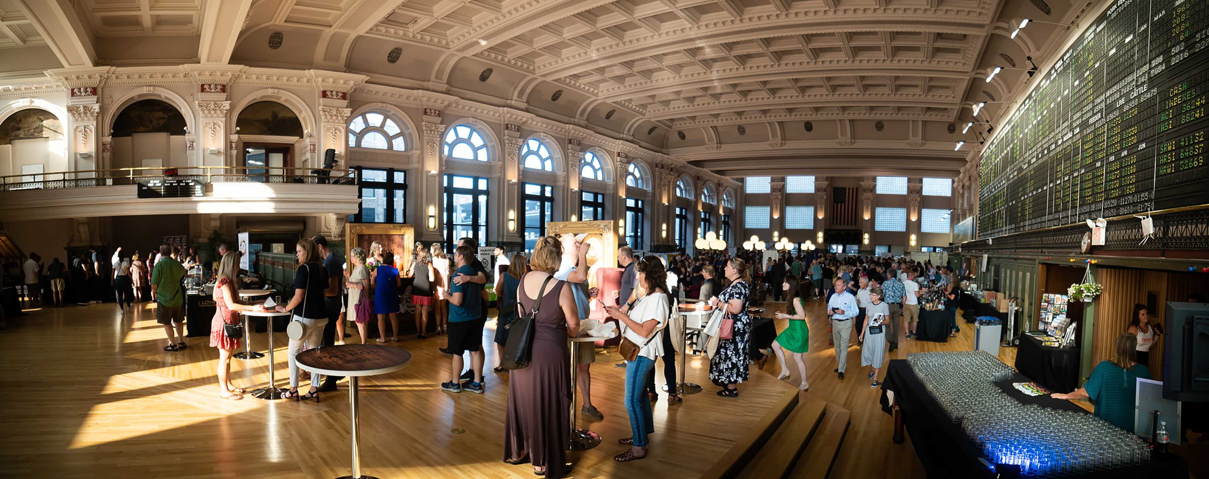 A large crowd gathers in an elegantly designed hall with high ceilings and tall windows, mingling at tables filled with food and drinks.