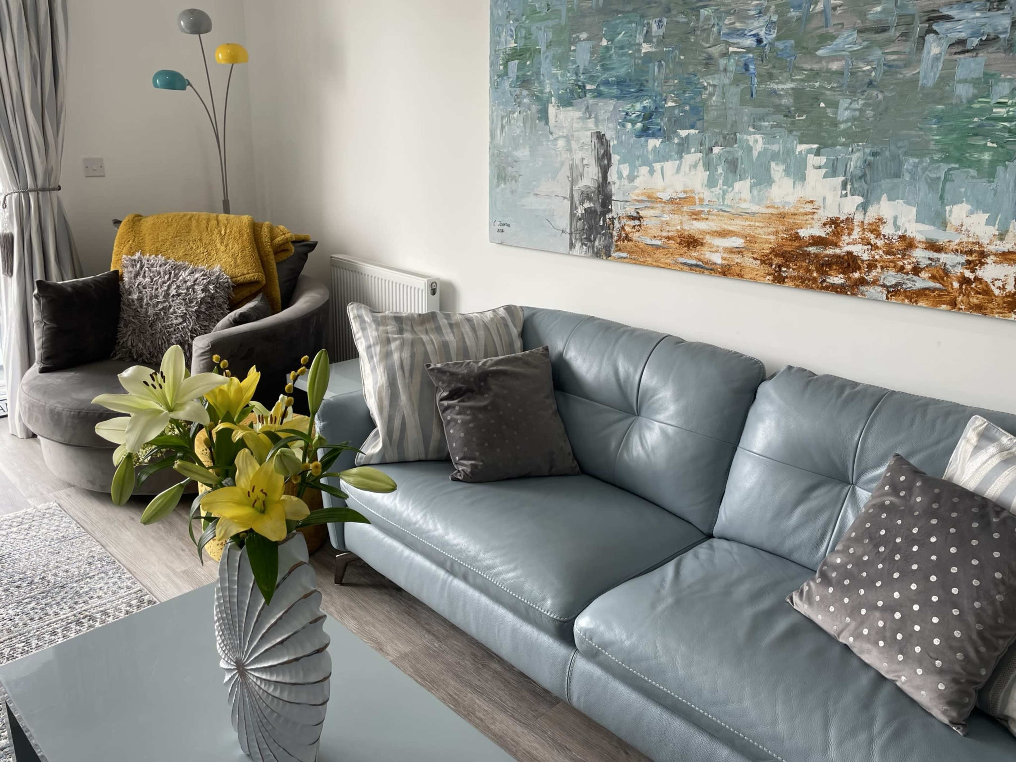 The image shows a modern living room featuring a light blue leather sofa, a coffee table with a decorative vase of flowers, and a large abstract painting on the wall.