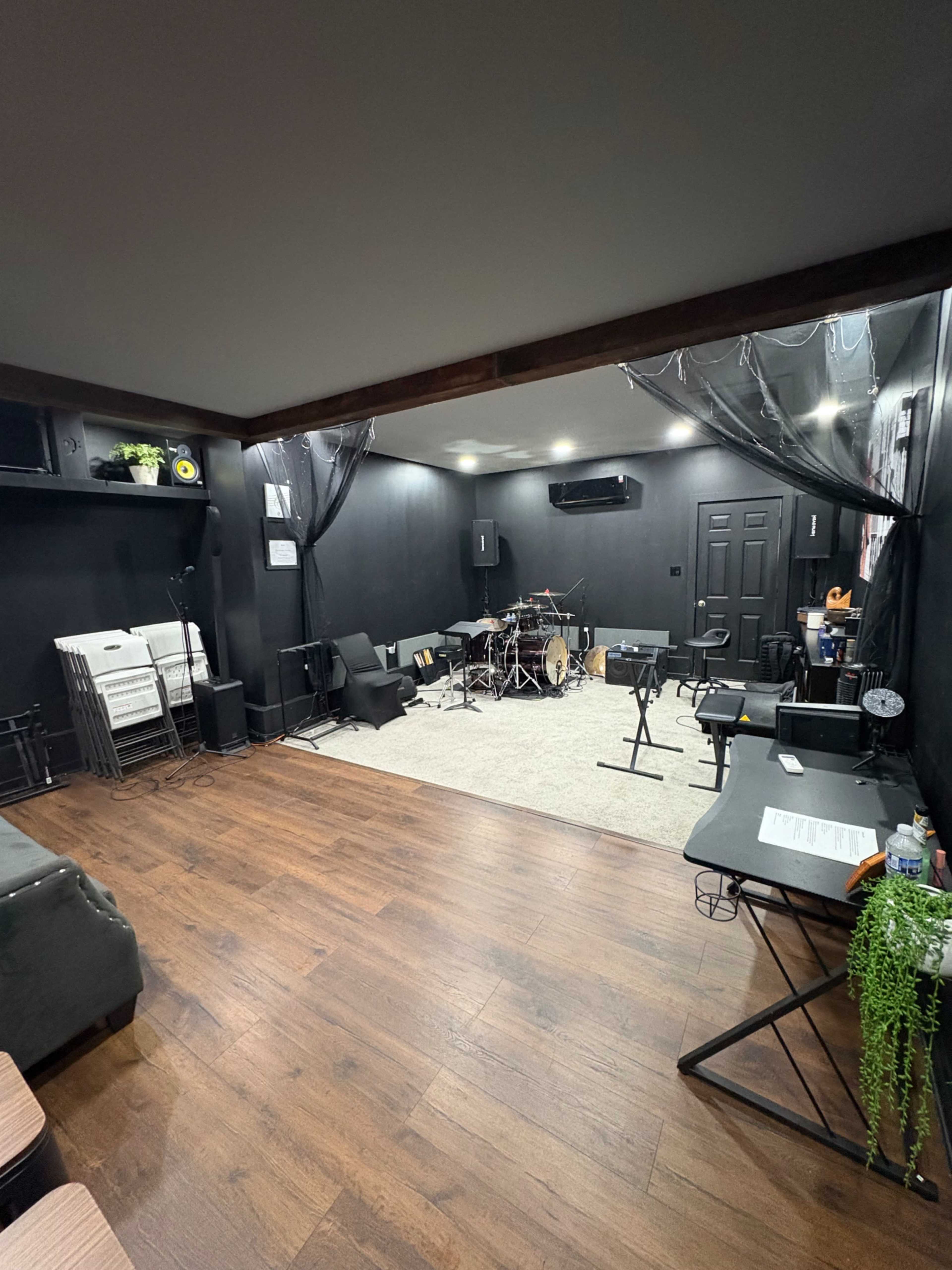 The image depicts a music rehearsal space with a drum set, various music stands, and seating arranged around a central area, all enclosed by black walls and a wooden floor.