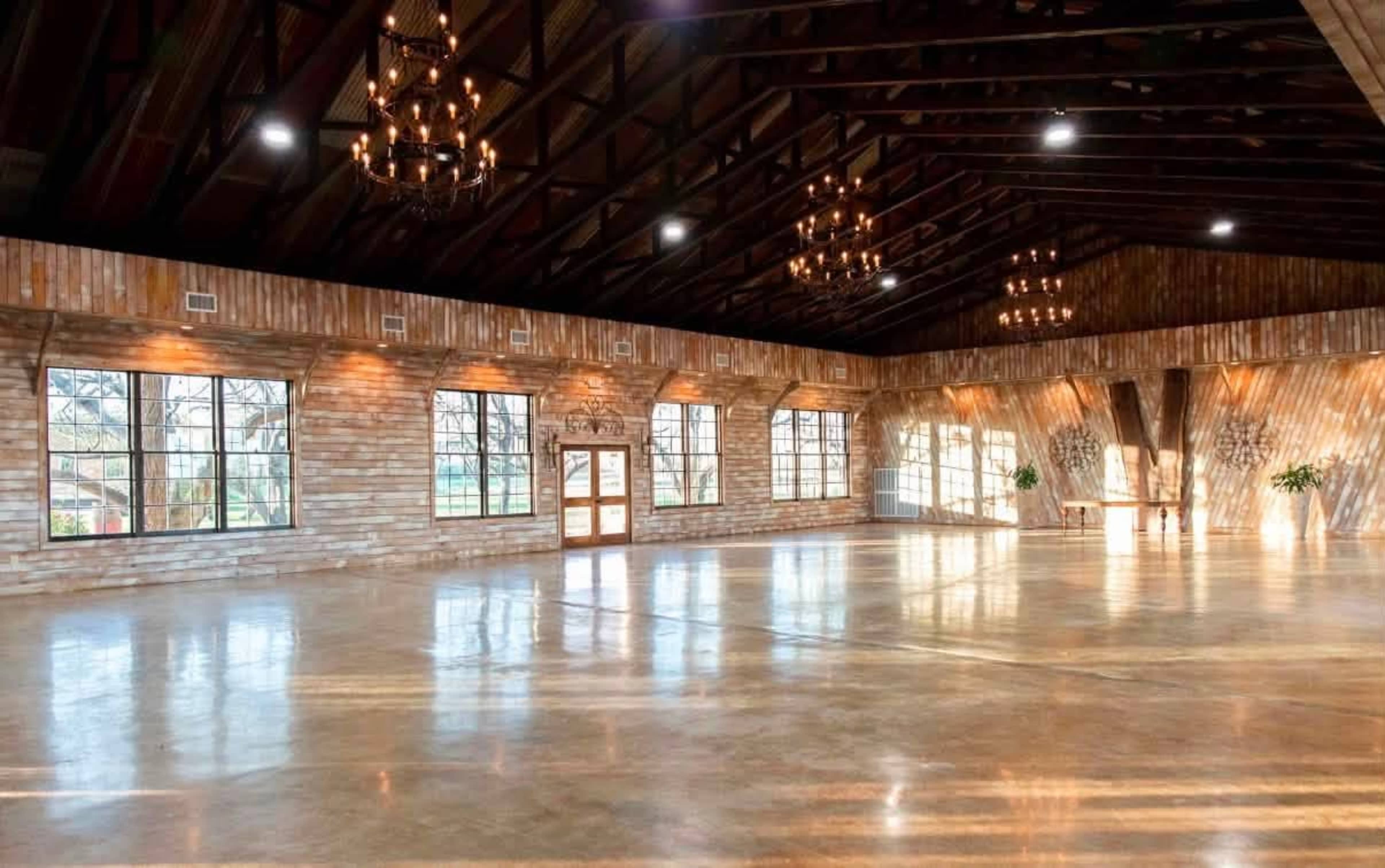 The image shows a spacious indoor venue with large windows, wooden walls, and chandeliers, featuring a polished concrete floor.
