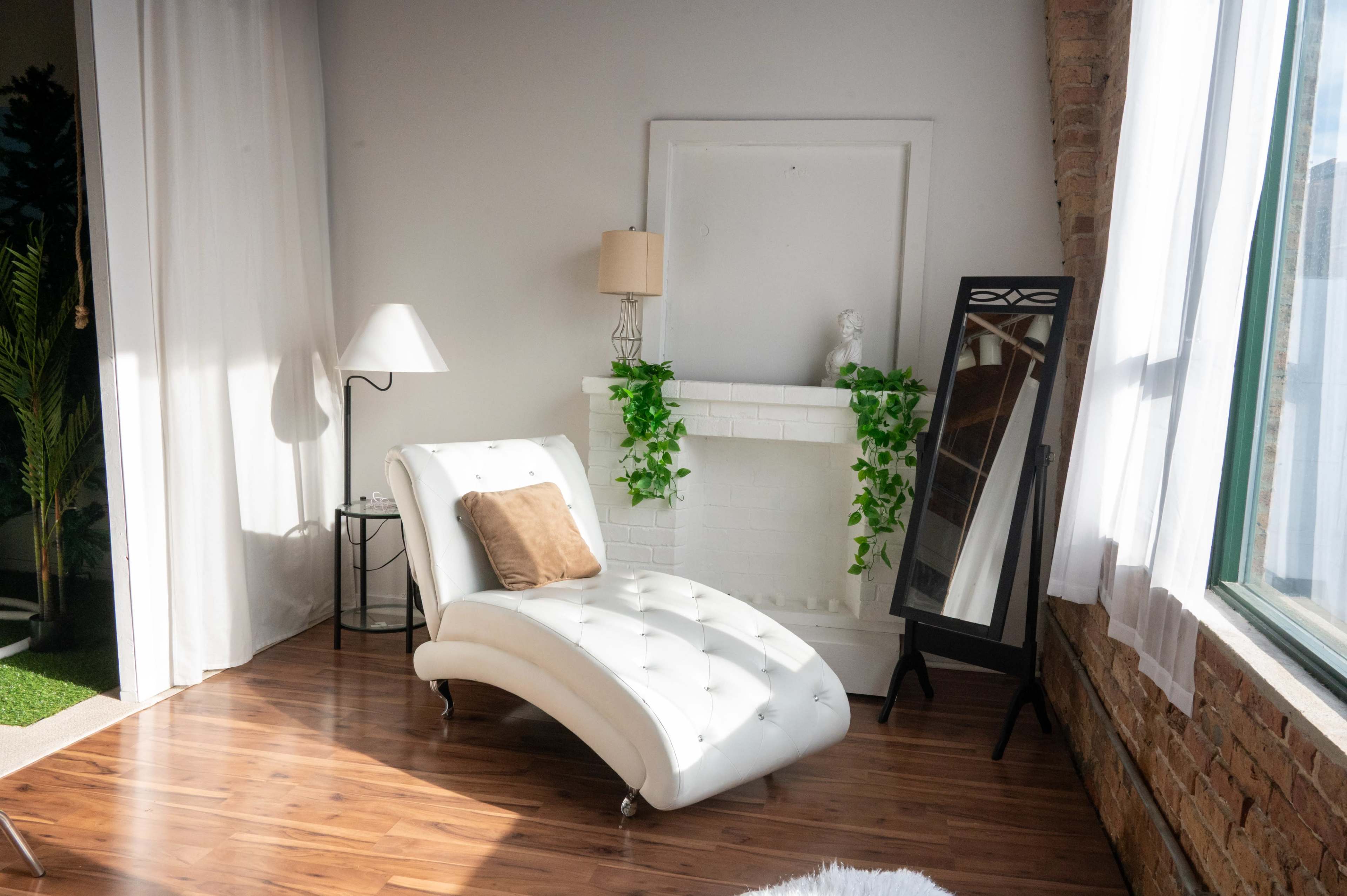 The image features a white chaise lounge next to a white fireplace adorned with greenery, a lamp, and a full-length mirror, all set in a well-lit room with wooden flooring and exposed brick walls.