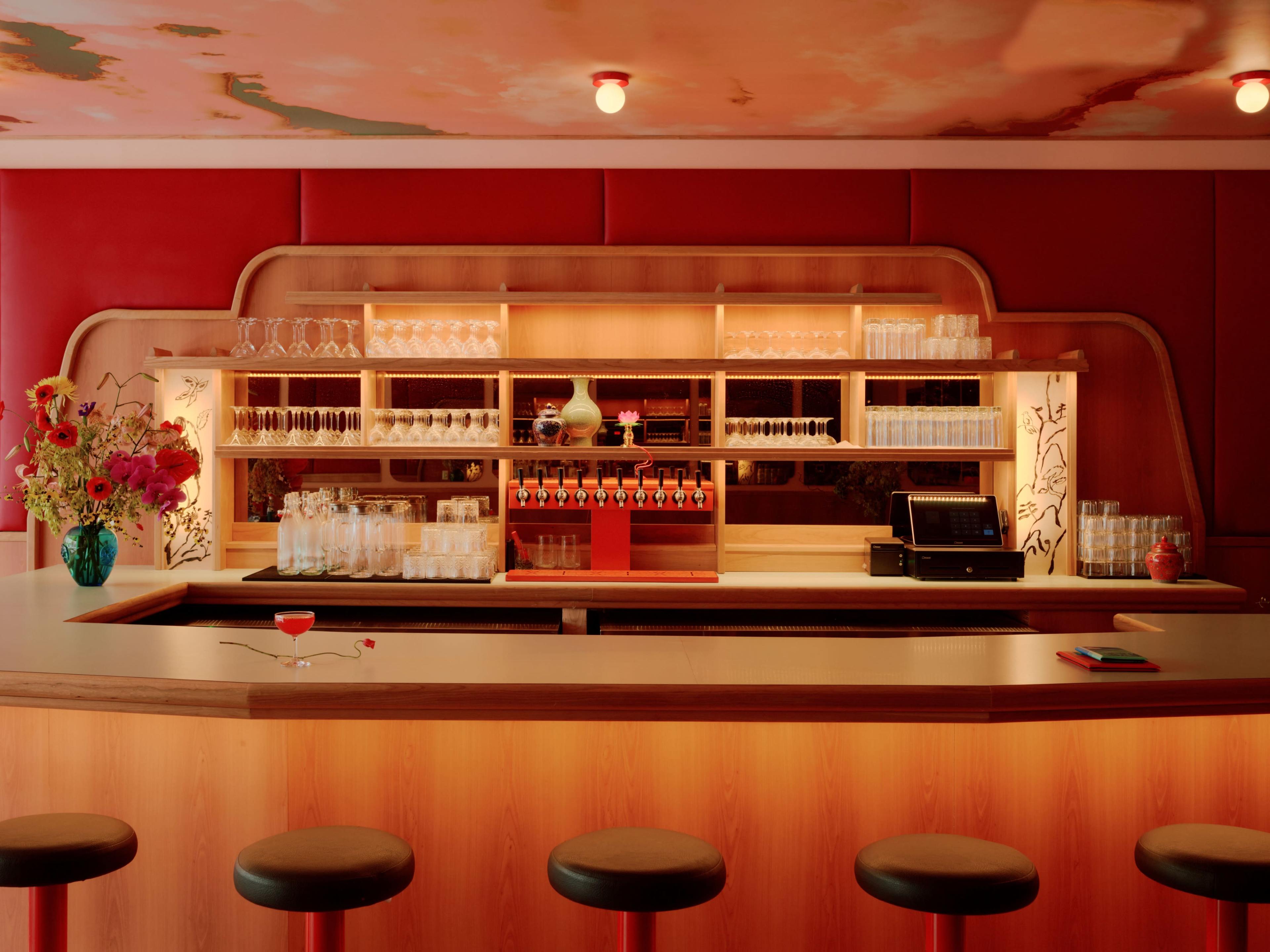 A bar featuring a wooden counter with stools, a backlit shelf displaying glasses and a beverage dispenser, and a floral arrangement on the counter.
