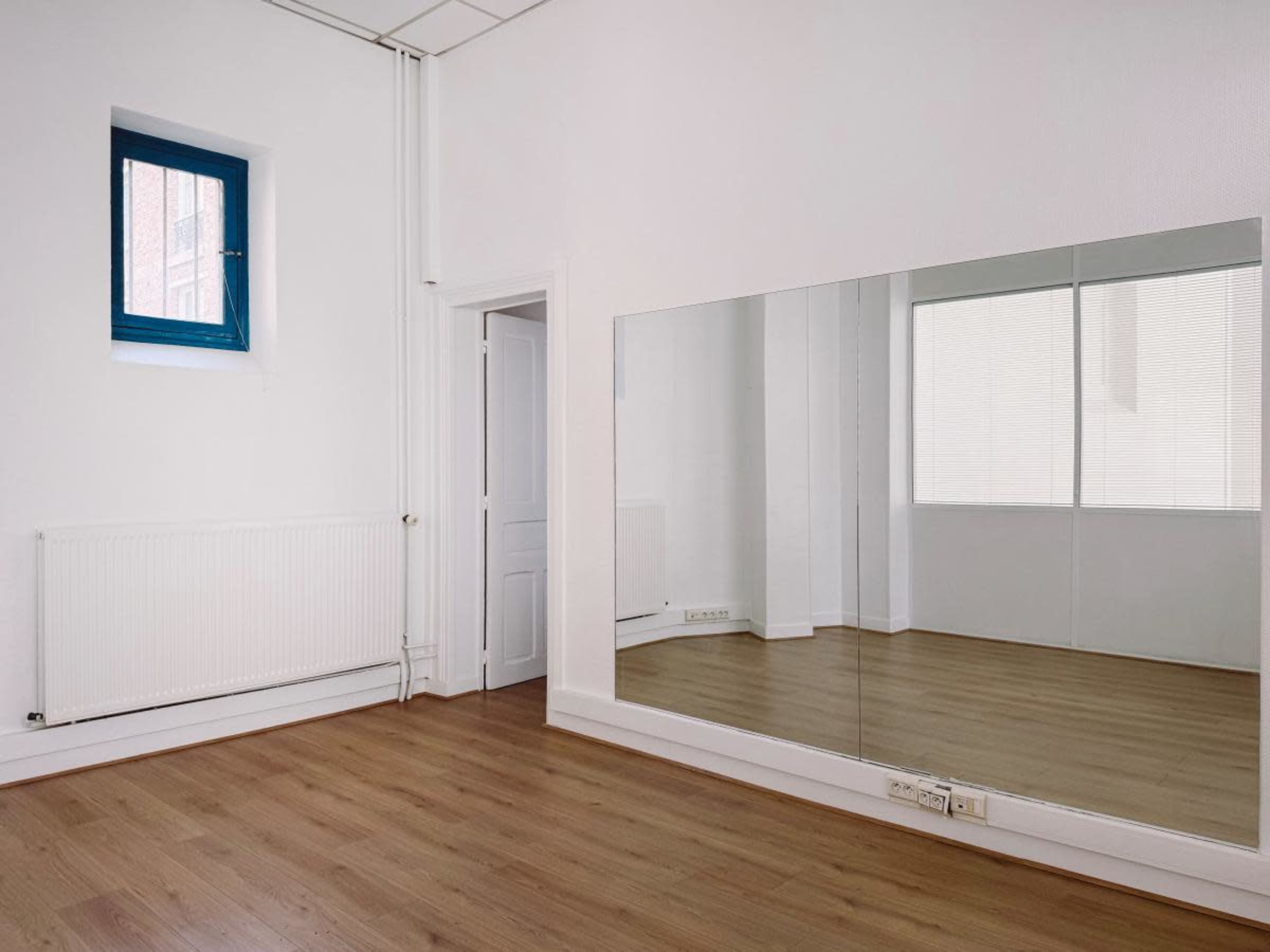 The image shows a sparsely furnished room with light-colored walls, a wooden floor, a rectangular mirror on one wall, and a blue window.