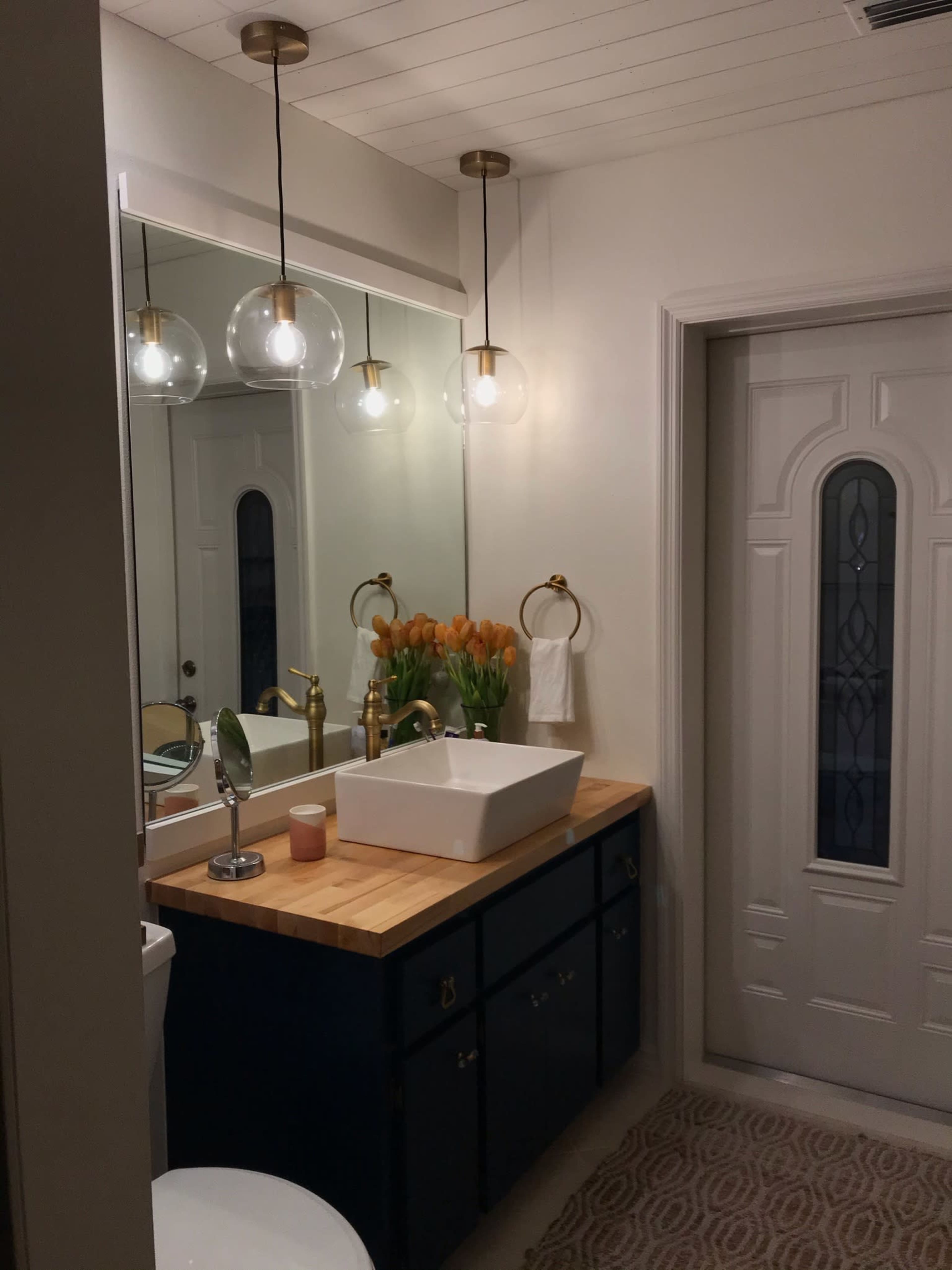 The image shows a modern bathroom with a wooden countertop, a white rectangular sink, a large mirror, and a vase of orange tulips, with a door leading to another room.