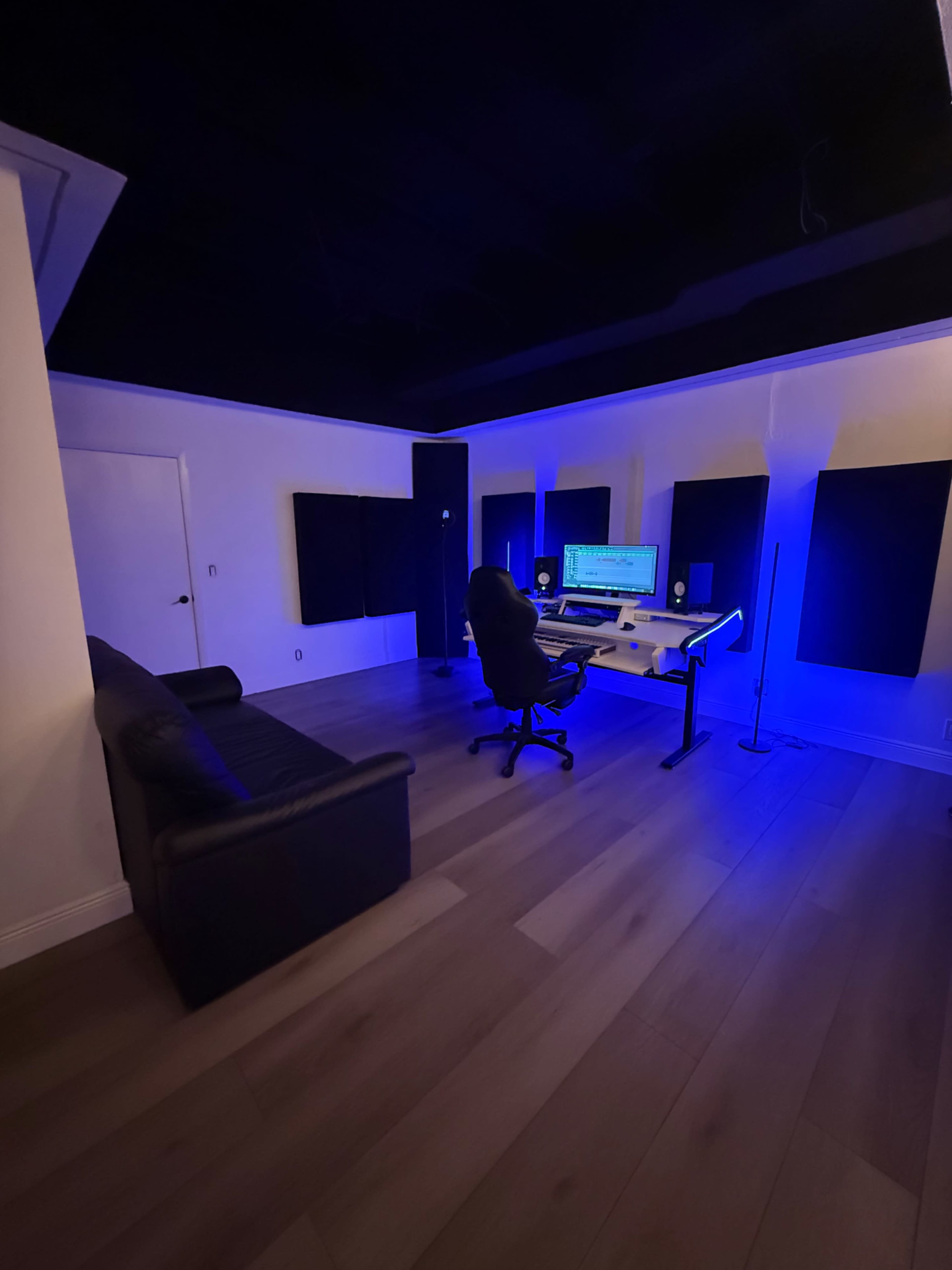 A music production studio features a desk with a computer, a rolling chair, acoustic panels on the walls, and ambient blue lighting.