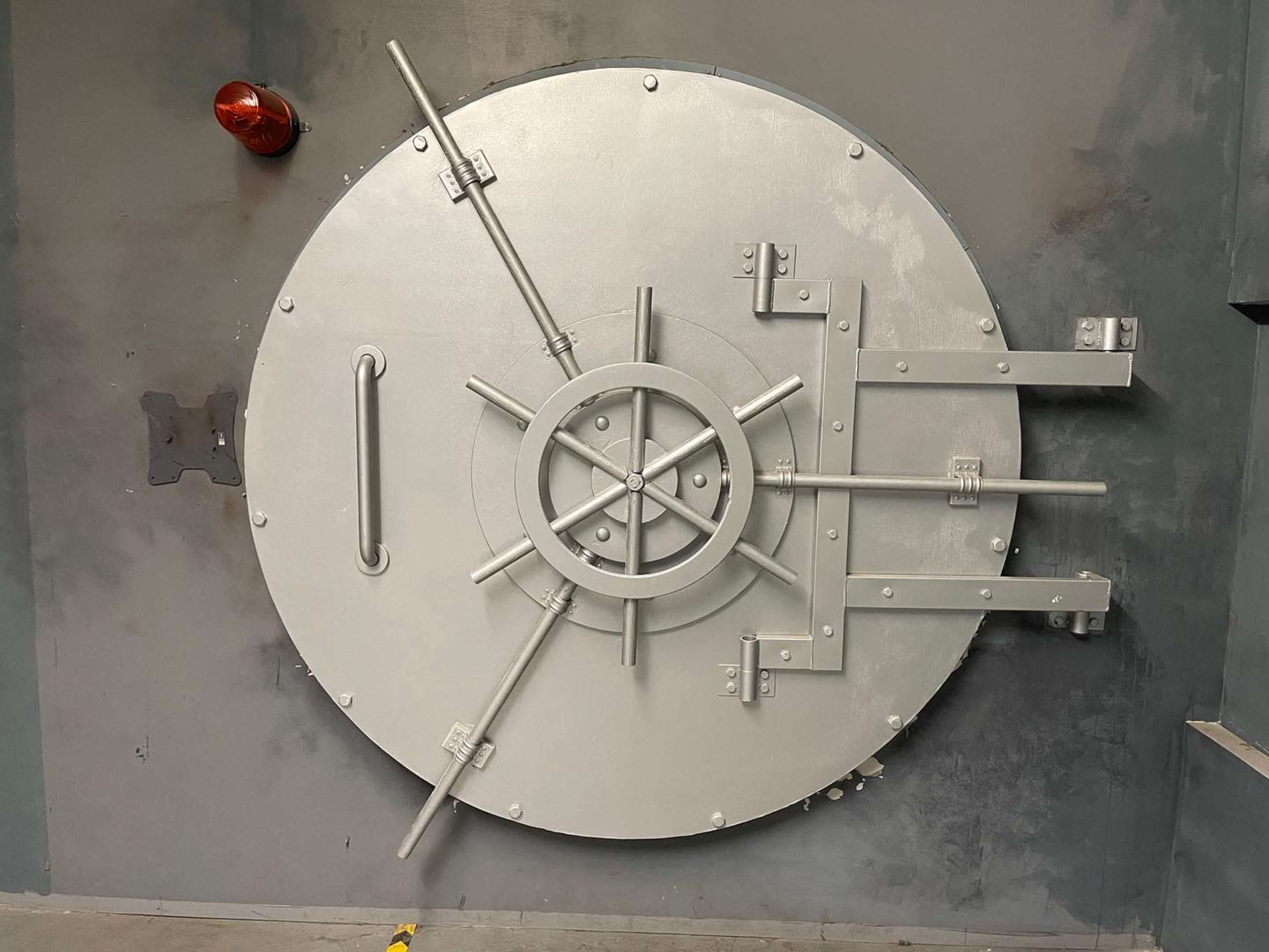 A large, metal vault door with a central locking mechanism and several bolts securing it against a gray wall.