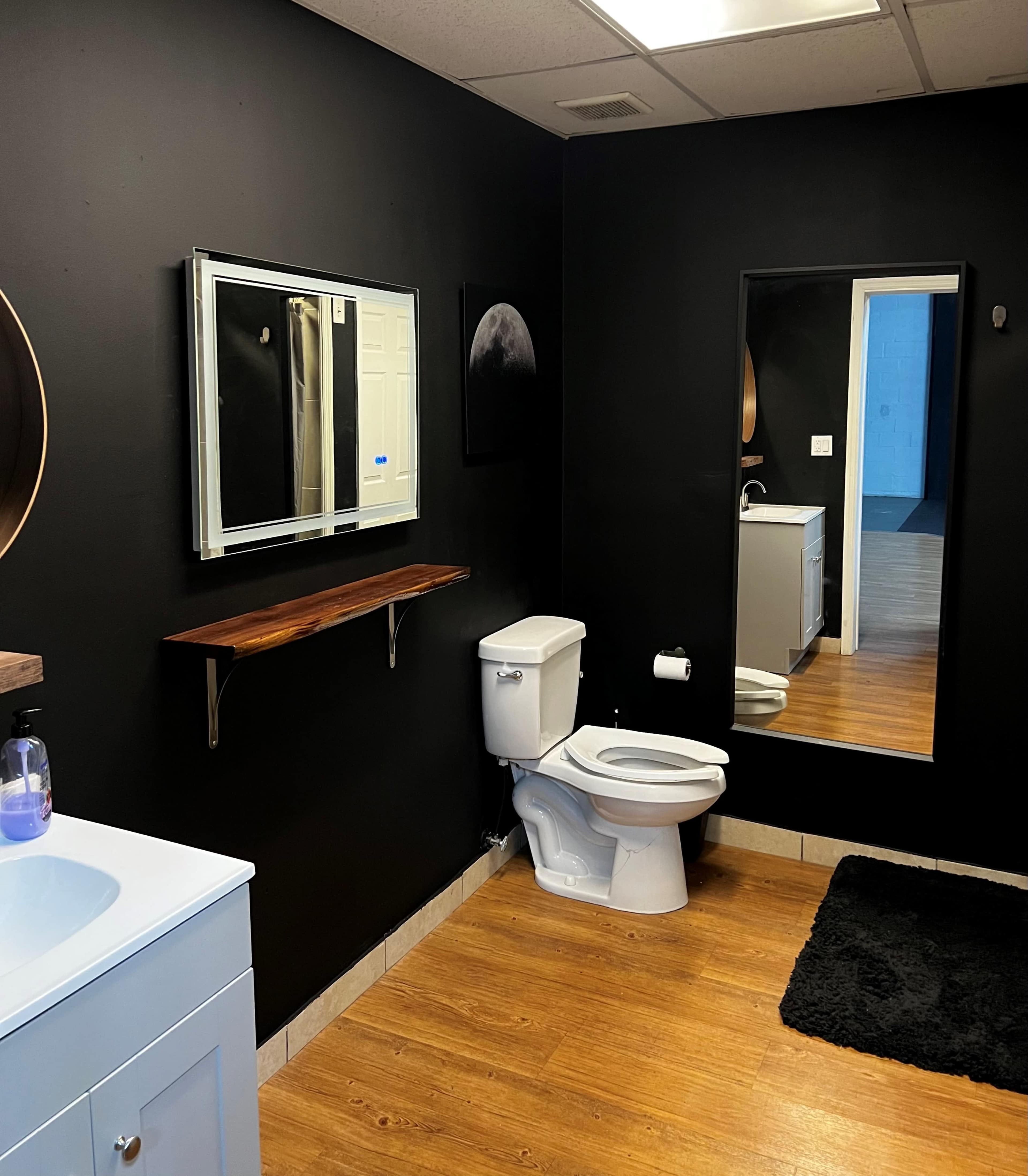 A modern bathroom with black walls, a white toilet, a mirror, and a wooden shelf.